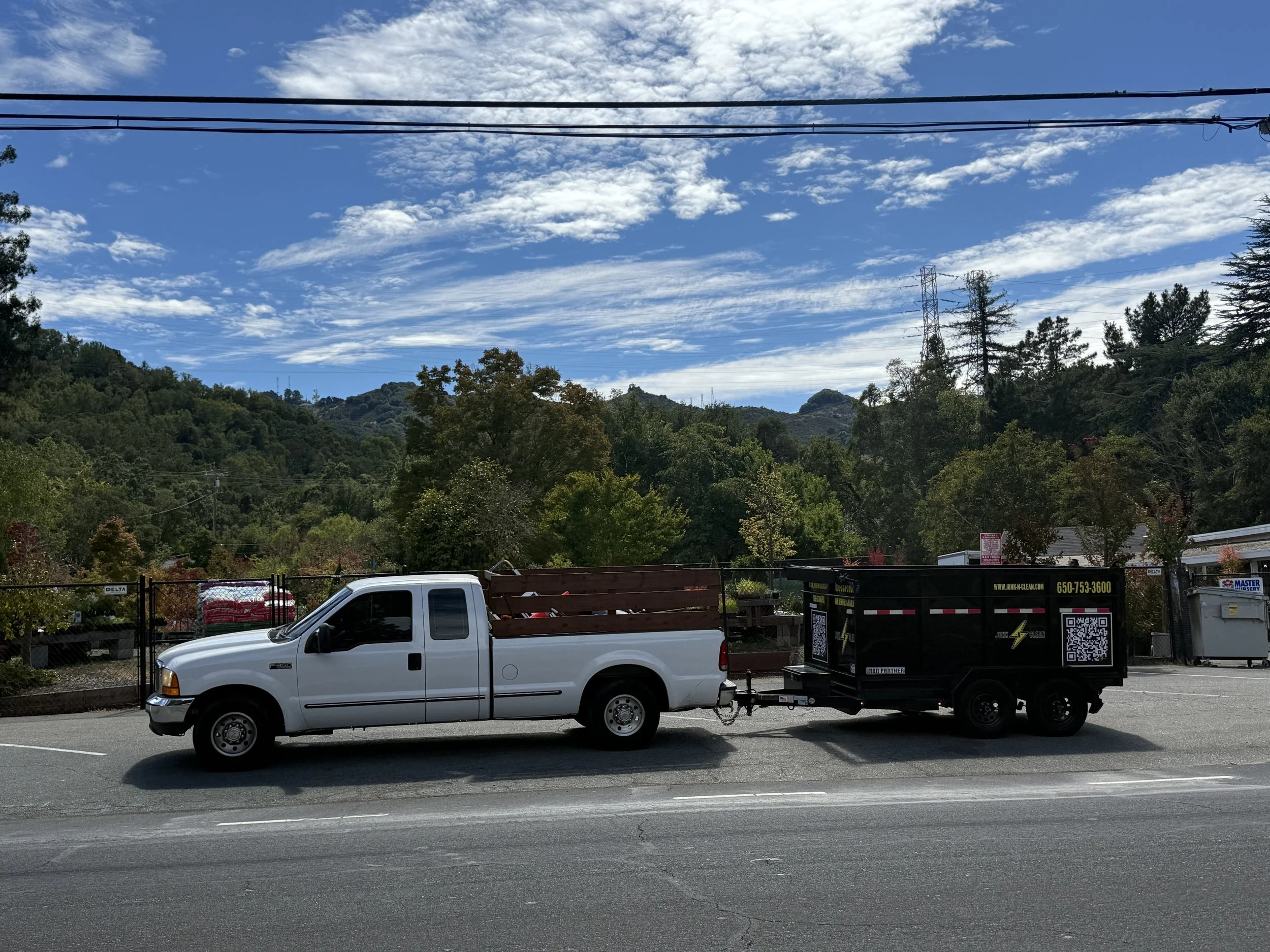 A white pickup truck connected to a black utility trailer is parked in a lot. In the background, there are trees, hills, power lines, and electrical towers under a partly cloudy sky.
