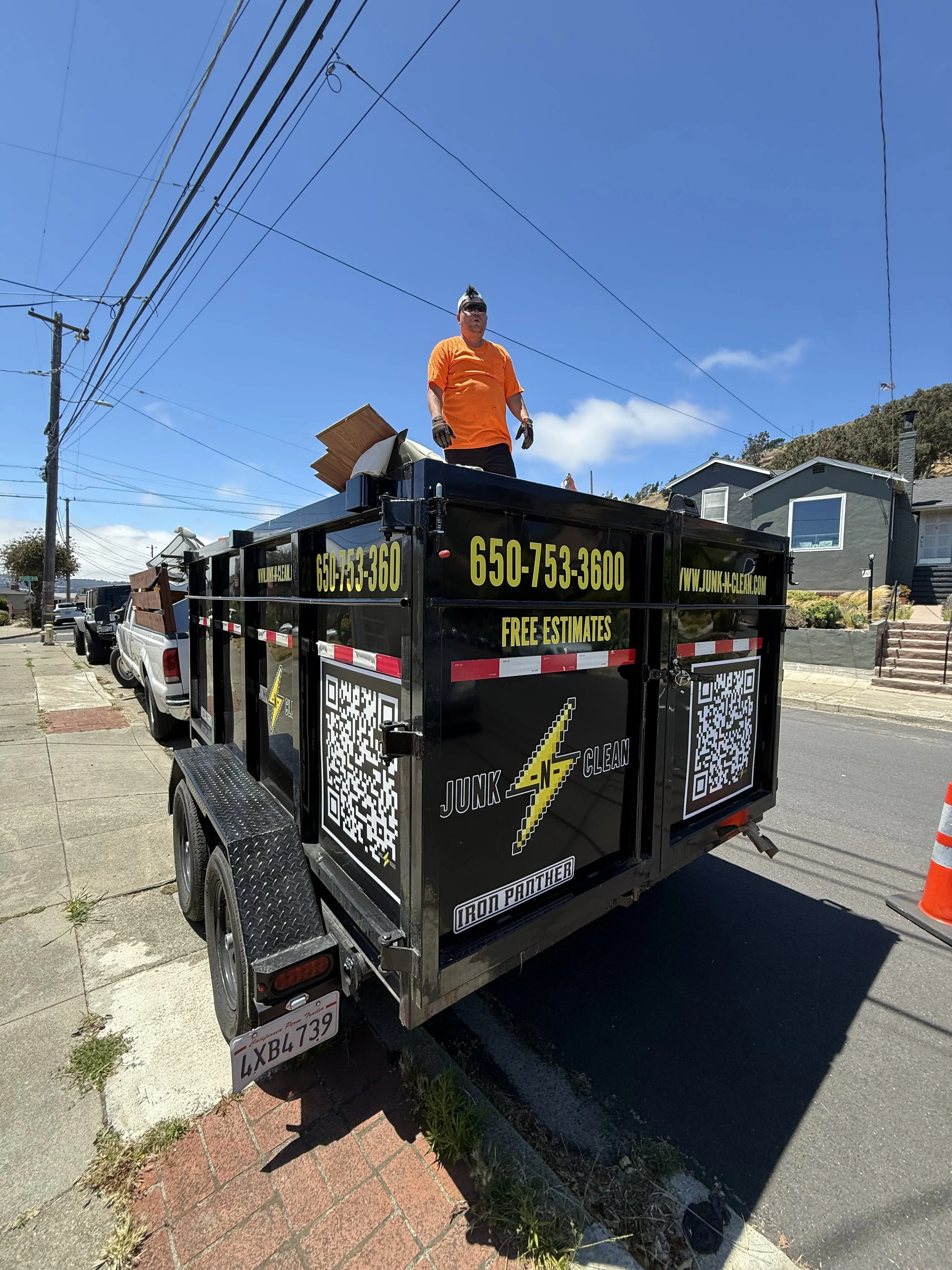 A man standing on a flatbed truck with the logo 'Junk N Clean' and contact information, parked on a residential street under a blue sky with houses and power lines in the background.