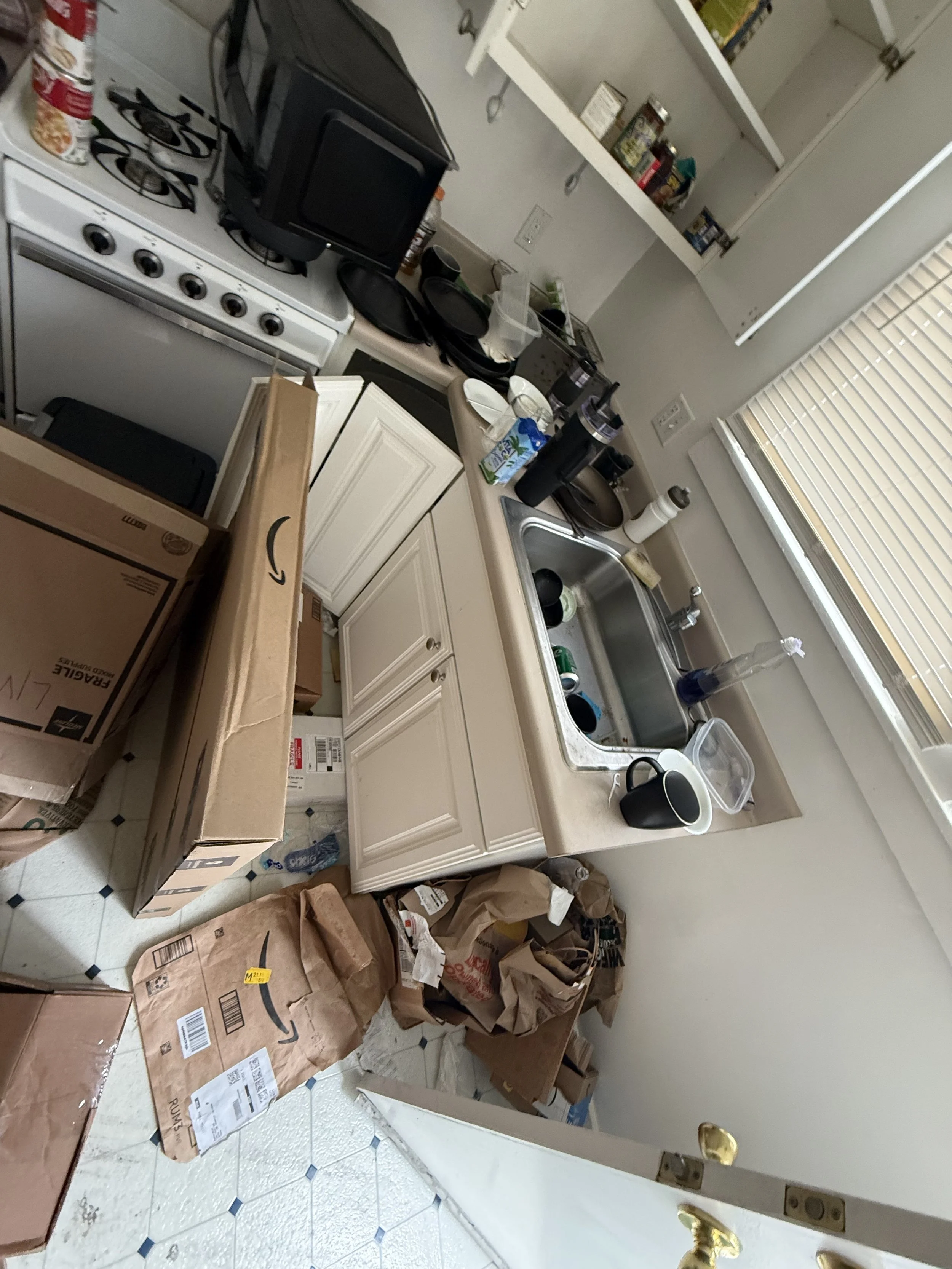 Cluttered kitchen with boxes and bags on the floor, open cabinet with canned goods, a stove with some small appliances and dishes on the countertop, and a window with blinds.