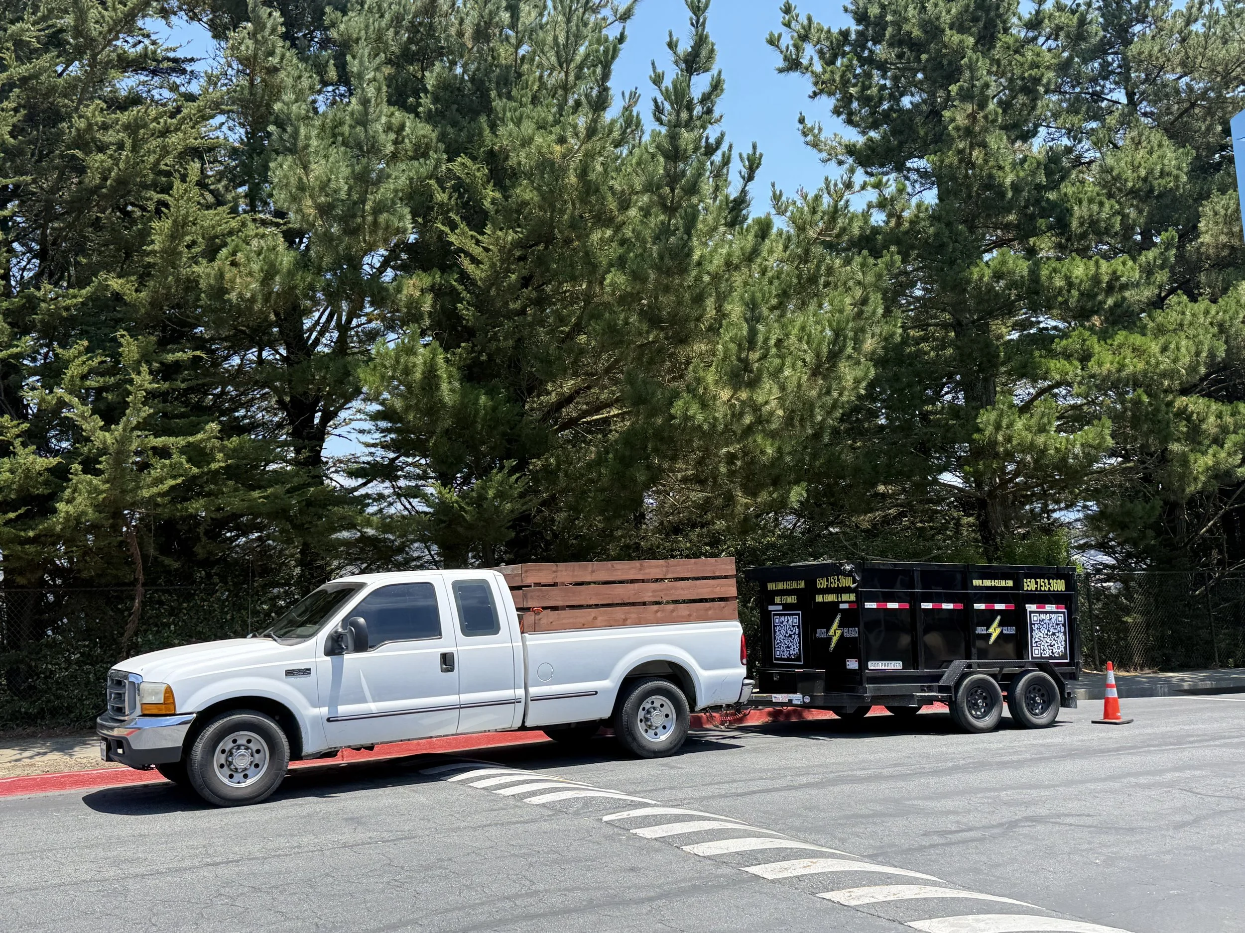 A white pickup truck with a trailer attached, parked along a street, with trees and a partly cloudy sky in the background.