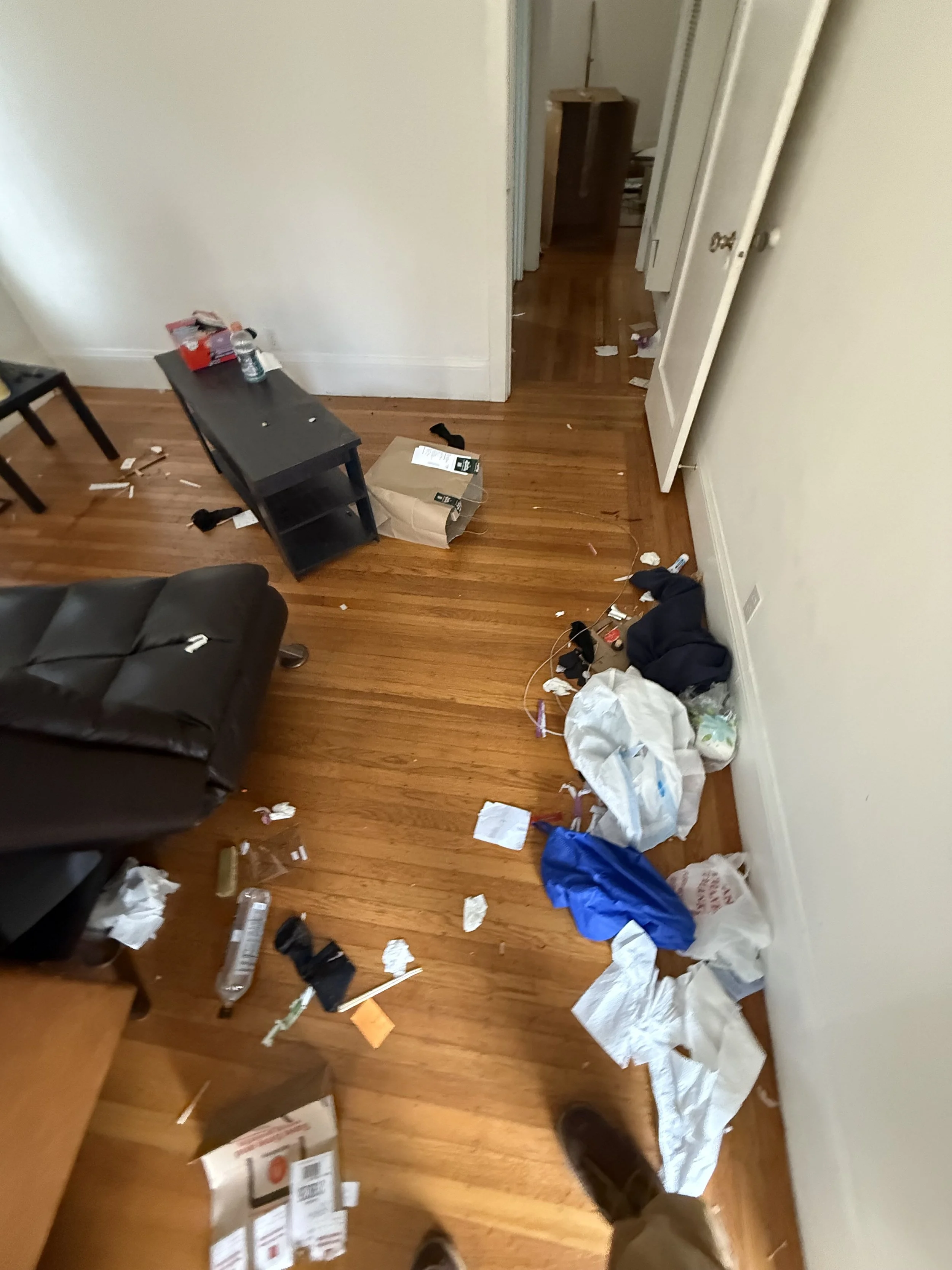 A cluttered room with trash and debris scattered on a hardwood floor, including paper, plastic, and discarded items. The image shows part of a black couch, a small black table, and a doorway leading to another room.