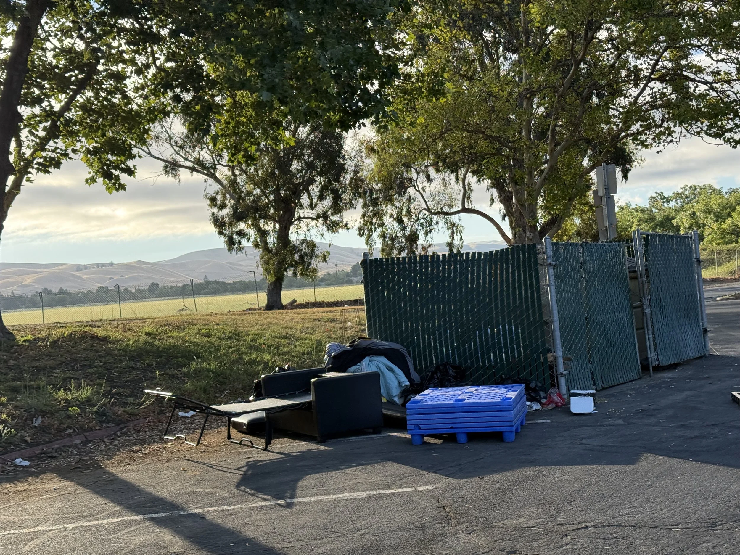 A park scene with trees, a chain-link fence, and a pile of items including black chairs, blue crates, and scattered trash on the ground. The background features rolling hills and a cloudy sky.
