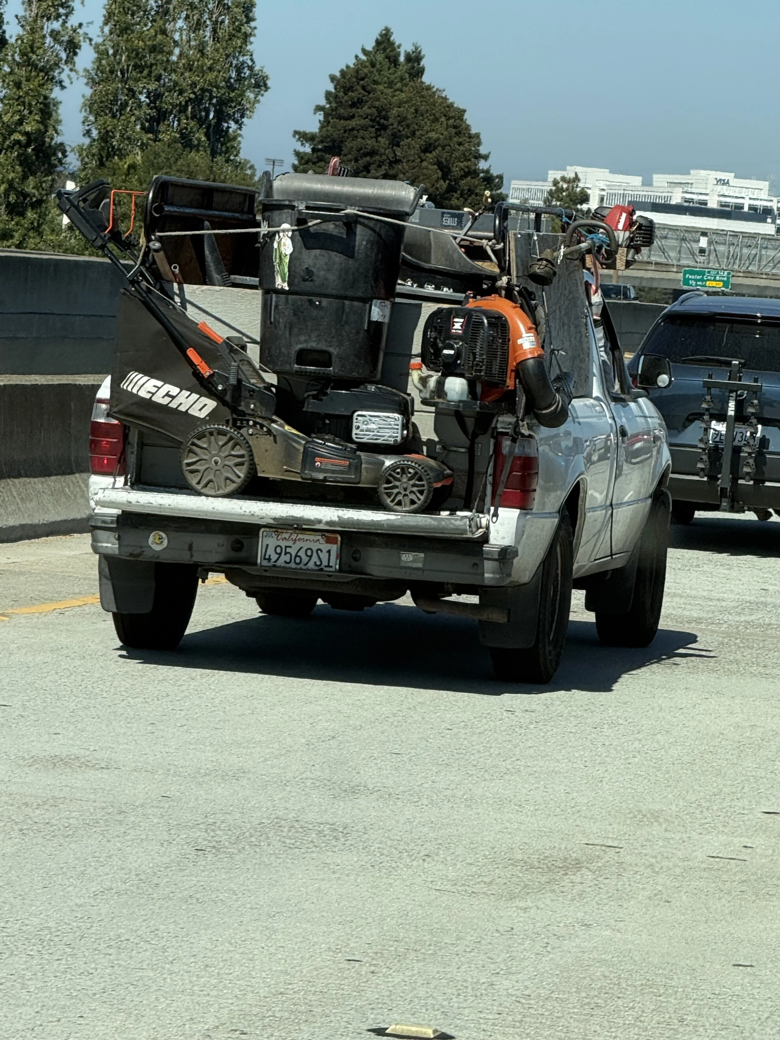 Silver pickup truck on highway carrying various over-sized equipment, including a lawn mower, a leaf blower, and other tools, with trees and a building in the background.