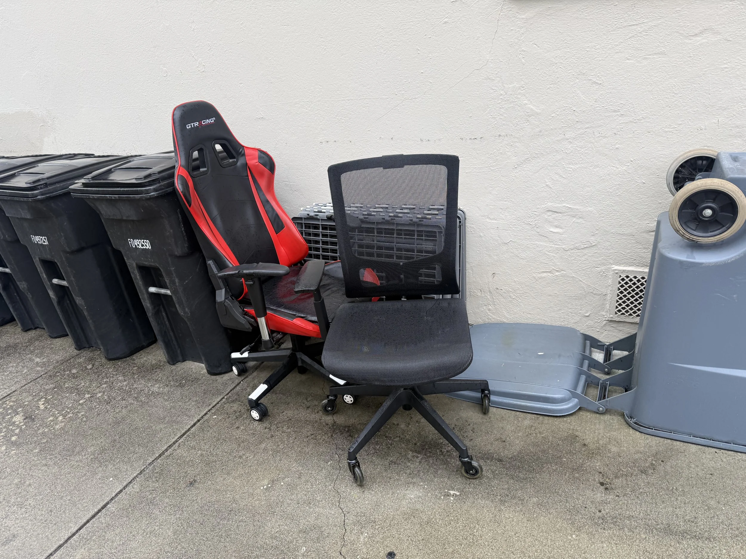 Various trash bins, an office chair, a racing seat, and other items are lined up against a white concrete wall outside on a sidewalk.