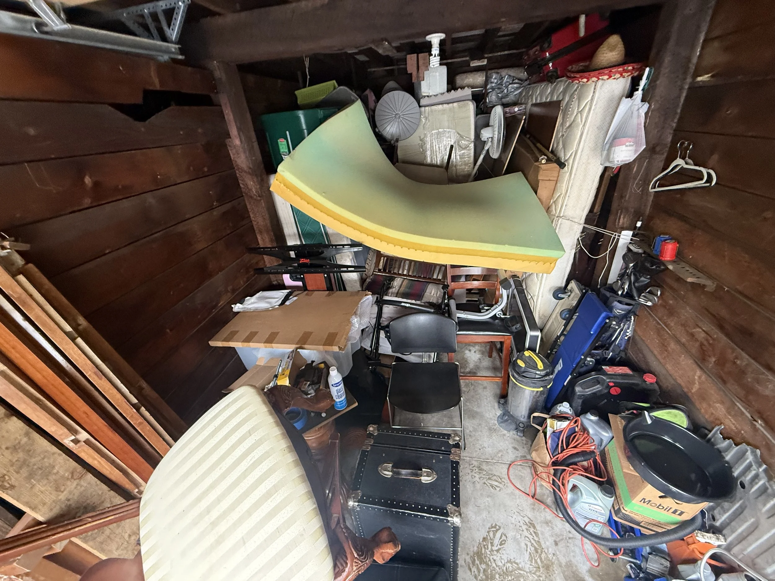 A cluttered storage room with wooden walls and a concrete floor, filled with various items including a foam mattress, chairs, a fan, a green plastic container, and various tools and boxes.