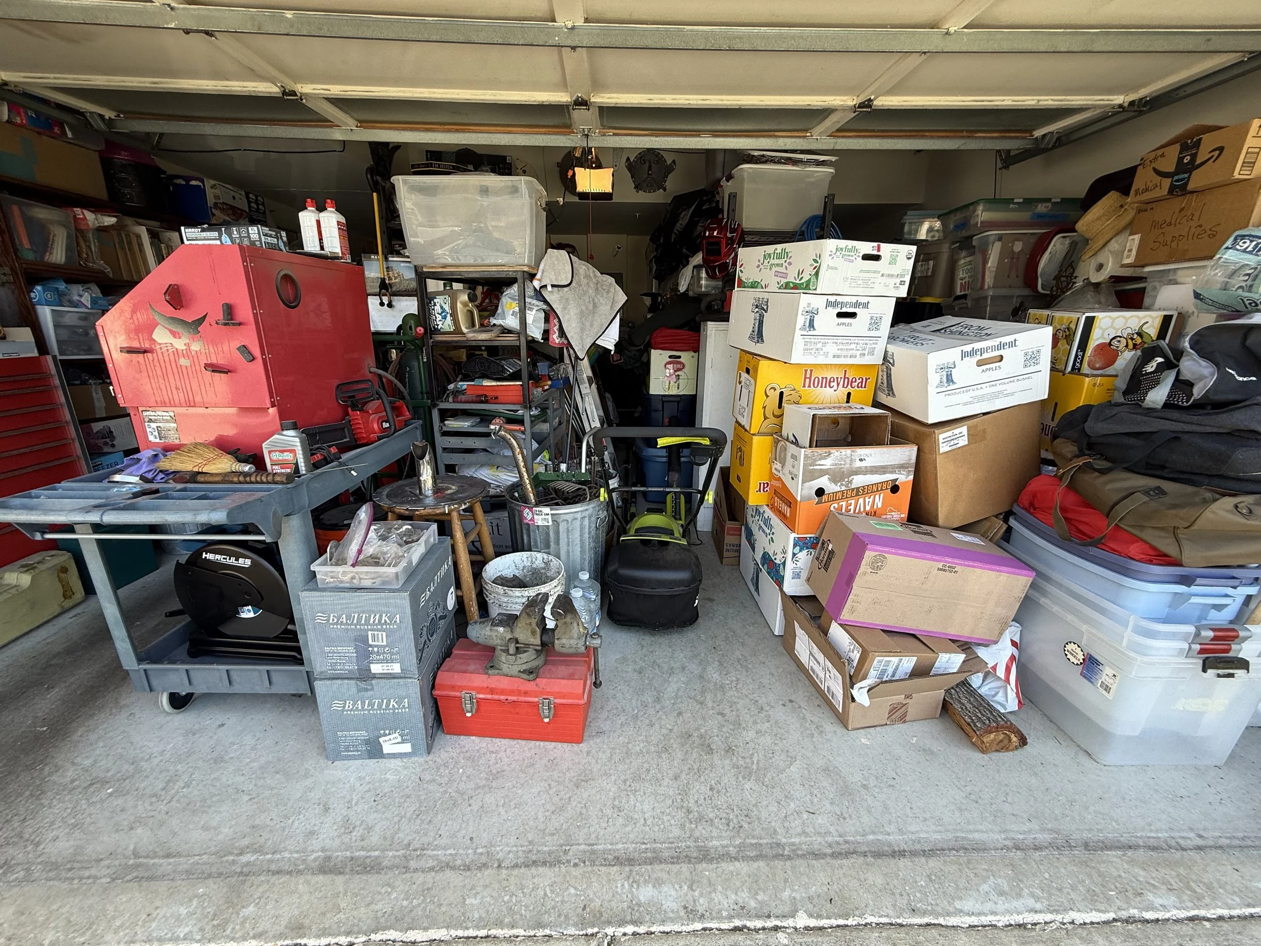 A cluttered garage filled with boxes, tools, and various household items, including a red tool chest, gas cans, boxes of produce, and shelves with supplies.