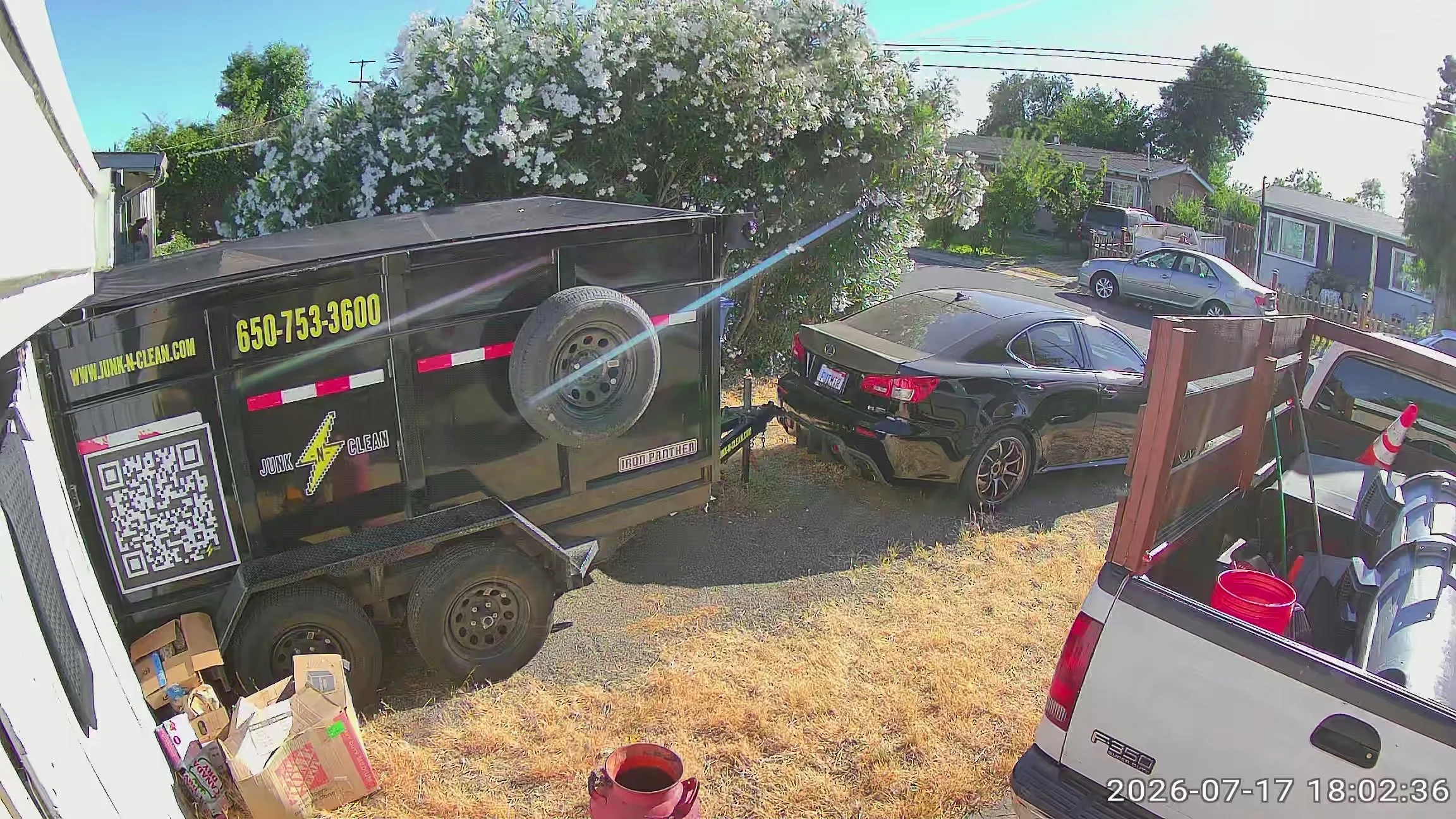 View of a residential backyard with a black car, a large black junk removal trailer, another white vehicle, and a hot tub. The yard has a large flowering bush and several trees, with neighboring houses visible in the background. Utility wires run acr