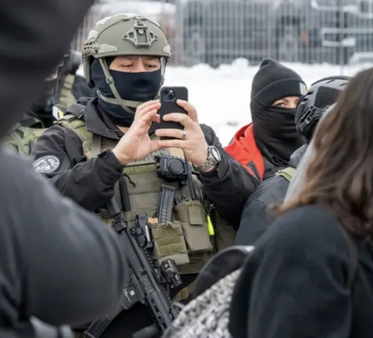 ICE agent in full gear and a face mask holding his phone up to focus on a protester's face