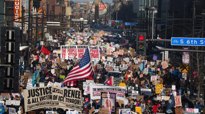 Looking down a street in Minneapolis filled sidewalk to sidewalk with marching people, holding signs that say "Justice for Renee & All Victims of ICE Terror", "The whole system is guilty!" and "Minnesota General Strike: The People Have the Power..."