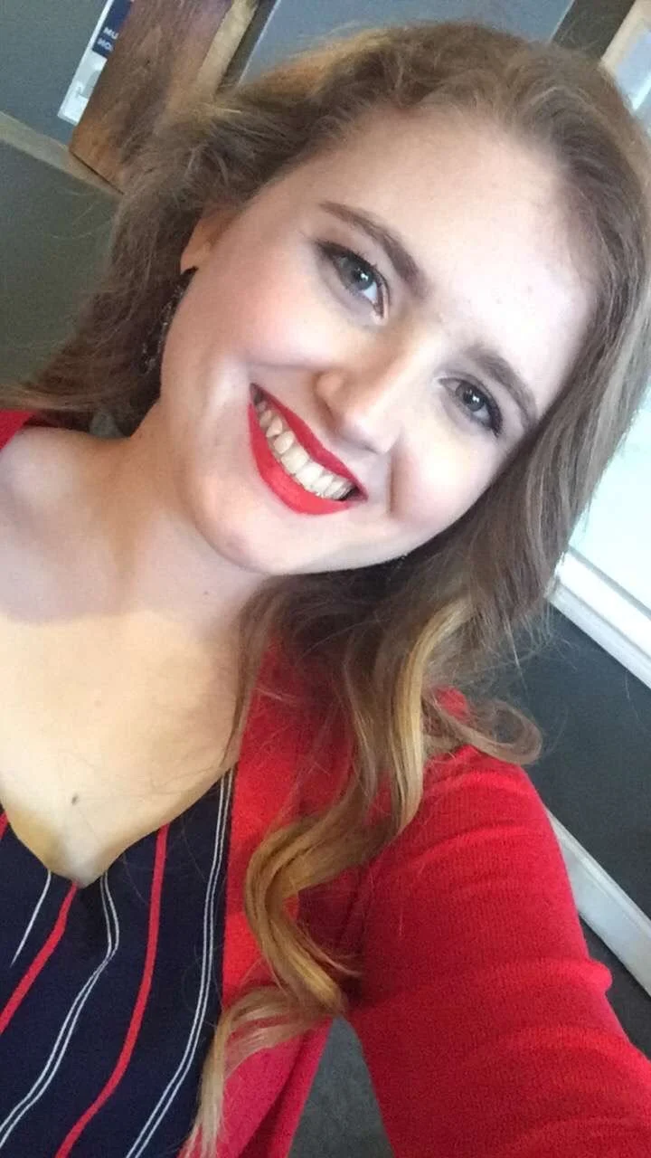A woman with long wavy brown hair, blue eyes, red lipstick, and a red and navy blue striped top smiling at the camera.