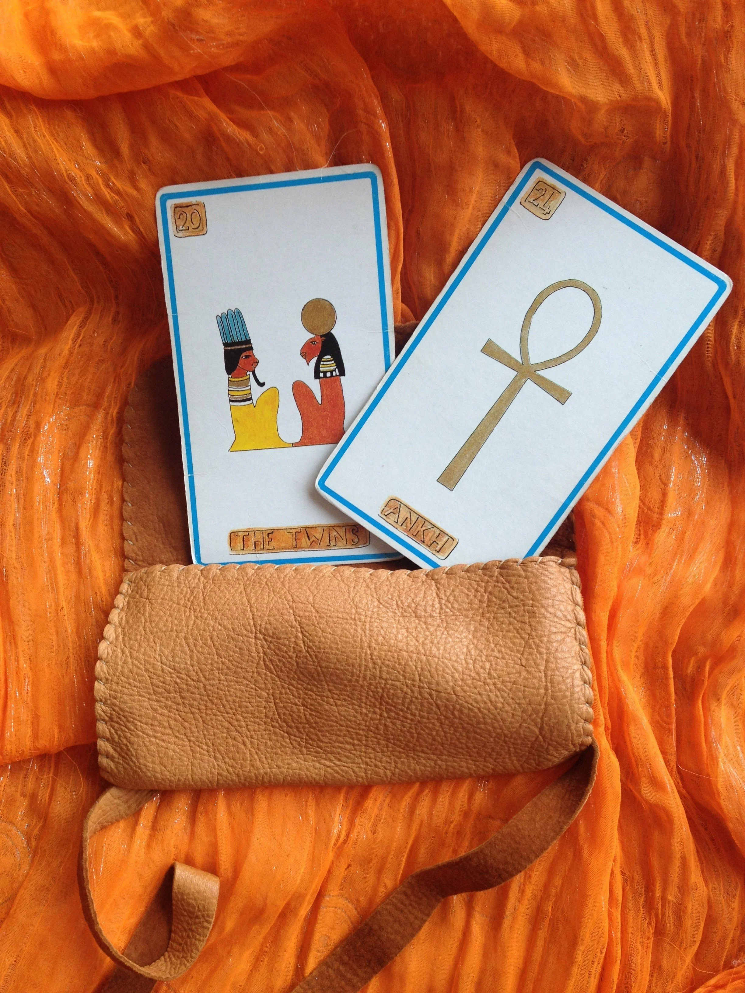 Two tarot cards, one labeled "The Twins" and the other with an ankh symbol, are placed on an orange textured fabric with a tan leather pouch beneath them.