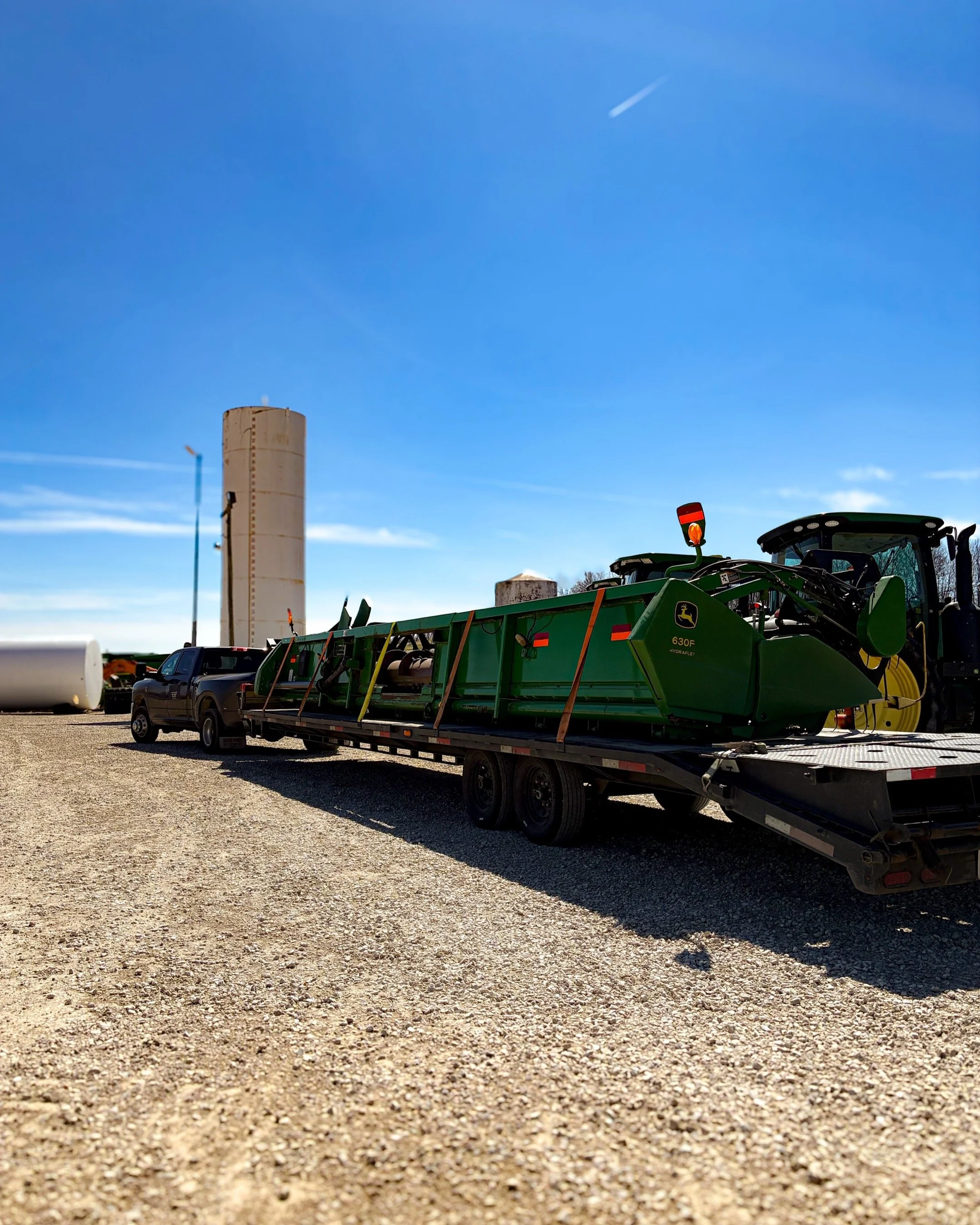 A flatbed trailer transporting farming equipment, including a John Deere 630F planter, on gravel ground with farm silos and machinery in the background under a clear blue sky.