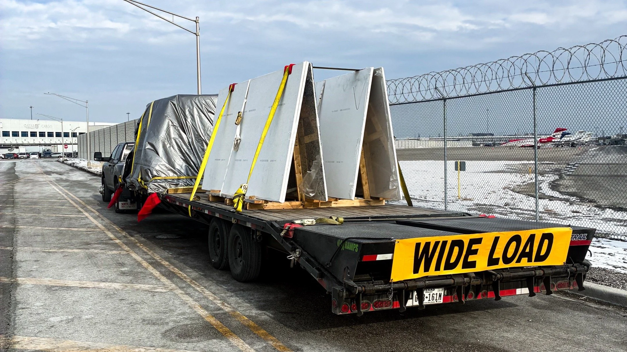 A flatbed truck with a yellow sign reading 'WIDE LOAD' carries large construction panels wrapped partially in black plastic near an airport tarmac with parked planes and barbed wire fence, during winter.