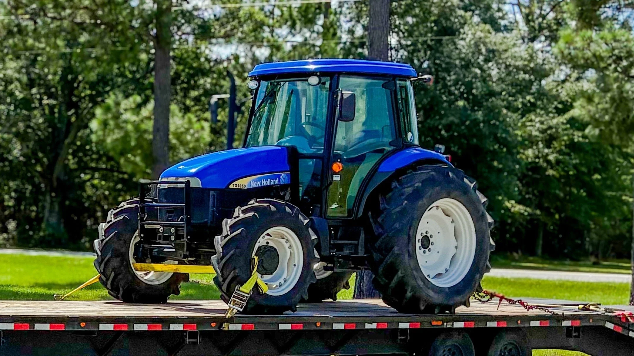 Blue New Holland tractor secured with straps on a flatbed trailer outdoors with green trees in the background.