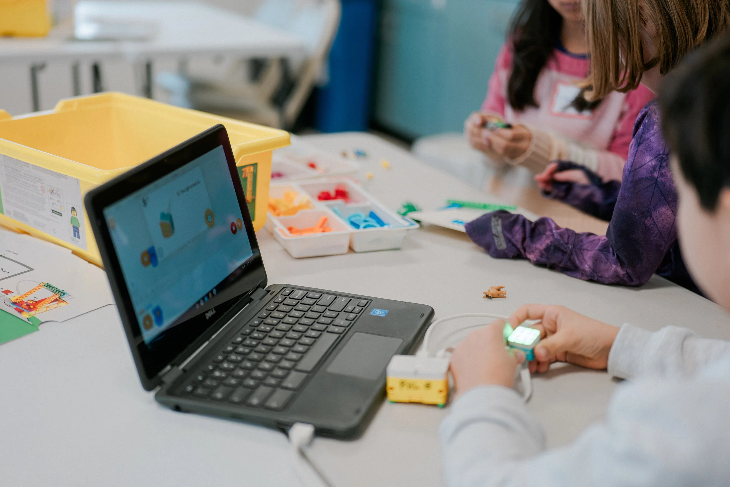 Children working on a robotics project with a small robot, LEGO parts, and a laptop on a table in a classroom.