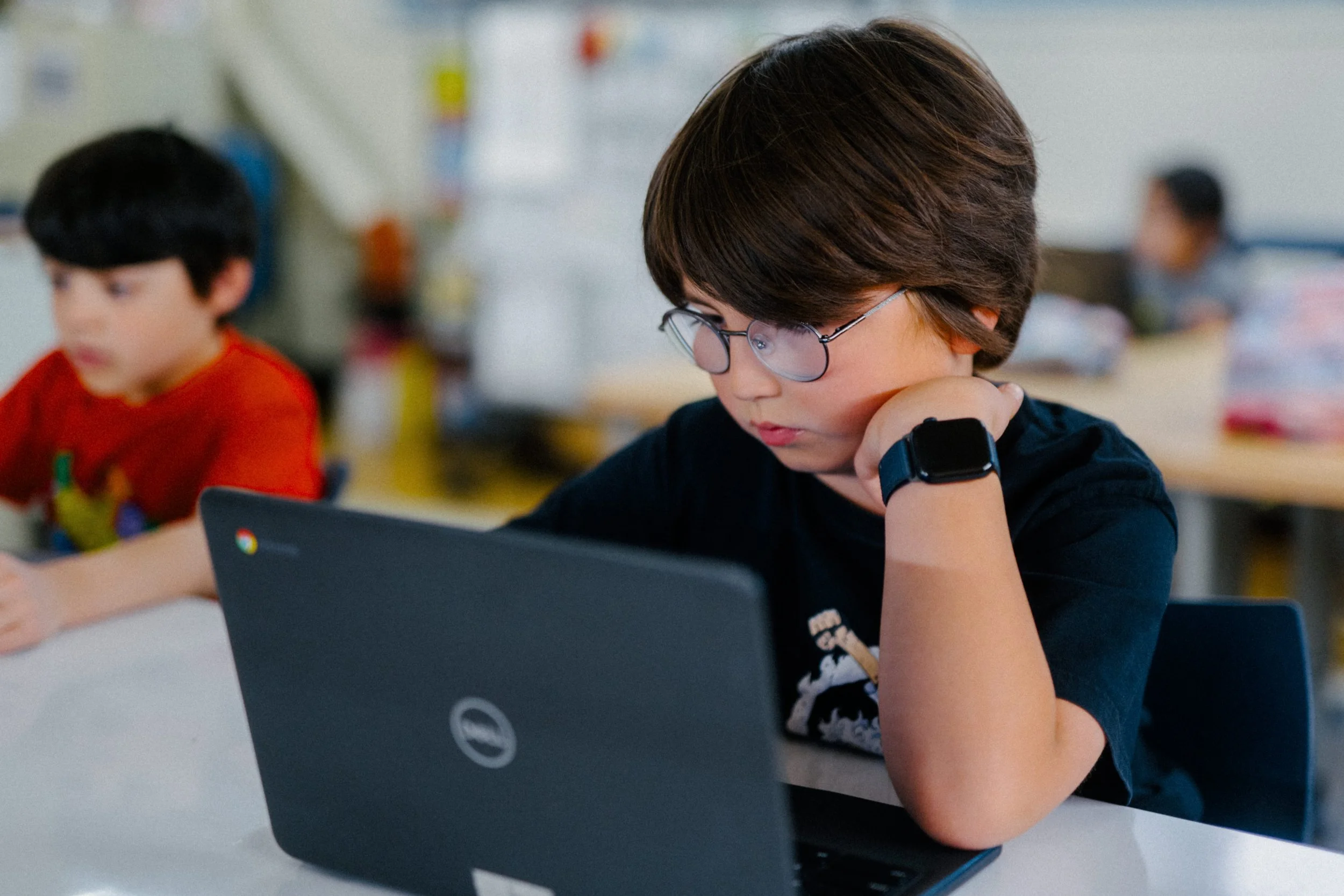 A young boy with glasses and a smartwatch sitting at a desk, focused on a laptop, with other children working in the background.