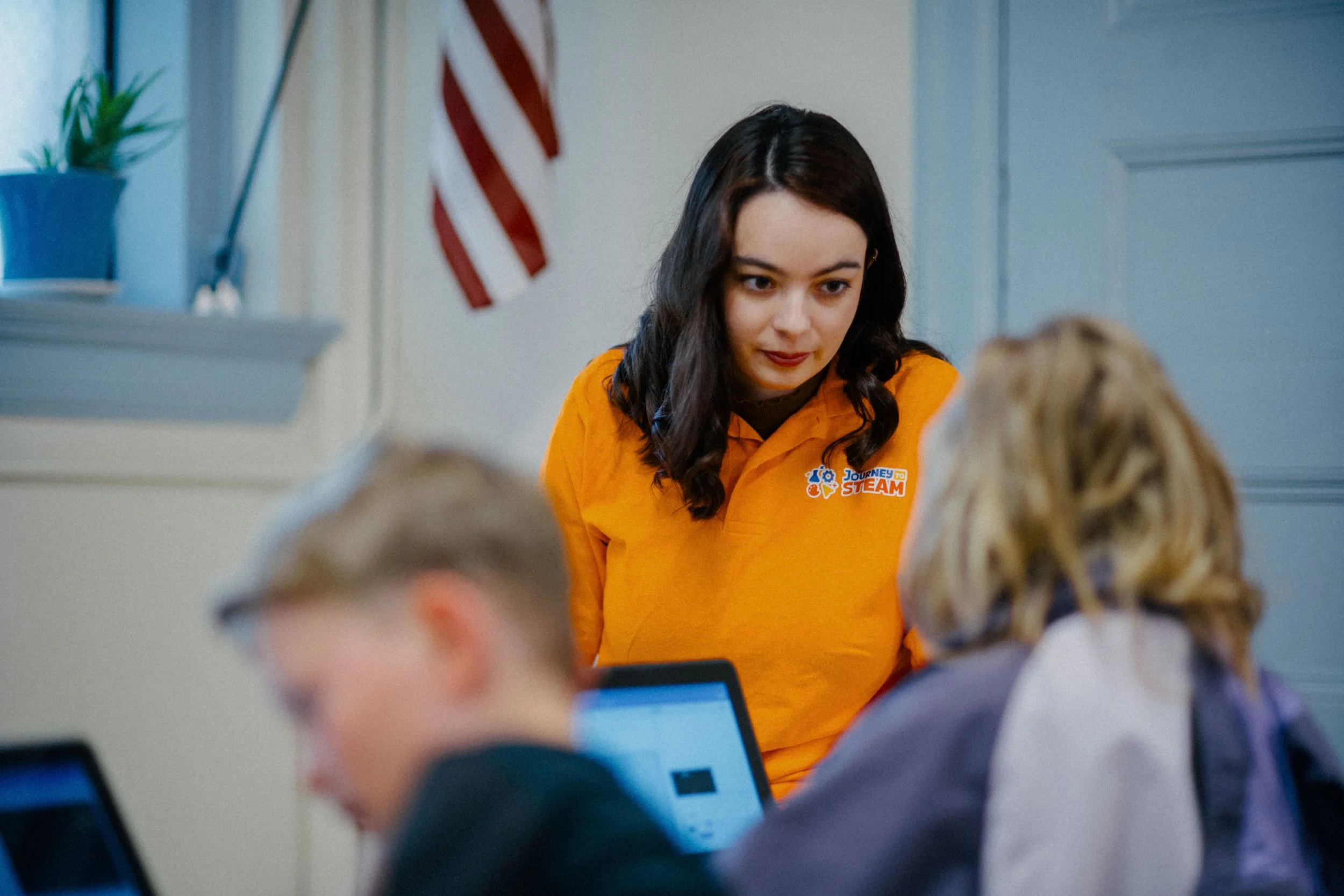 A young woman in an orange "Journey to STEAM" polo shirt is speaking to two students using laptops in a classroom, with an American flag and a potted plant in the background.