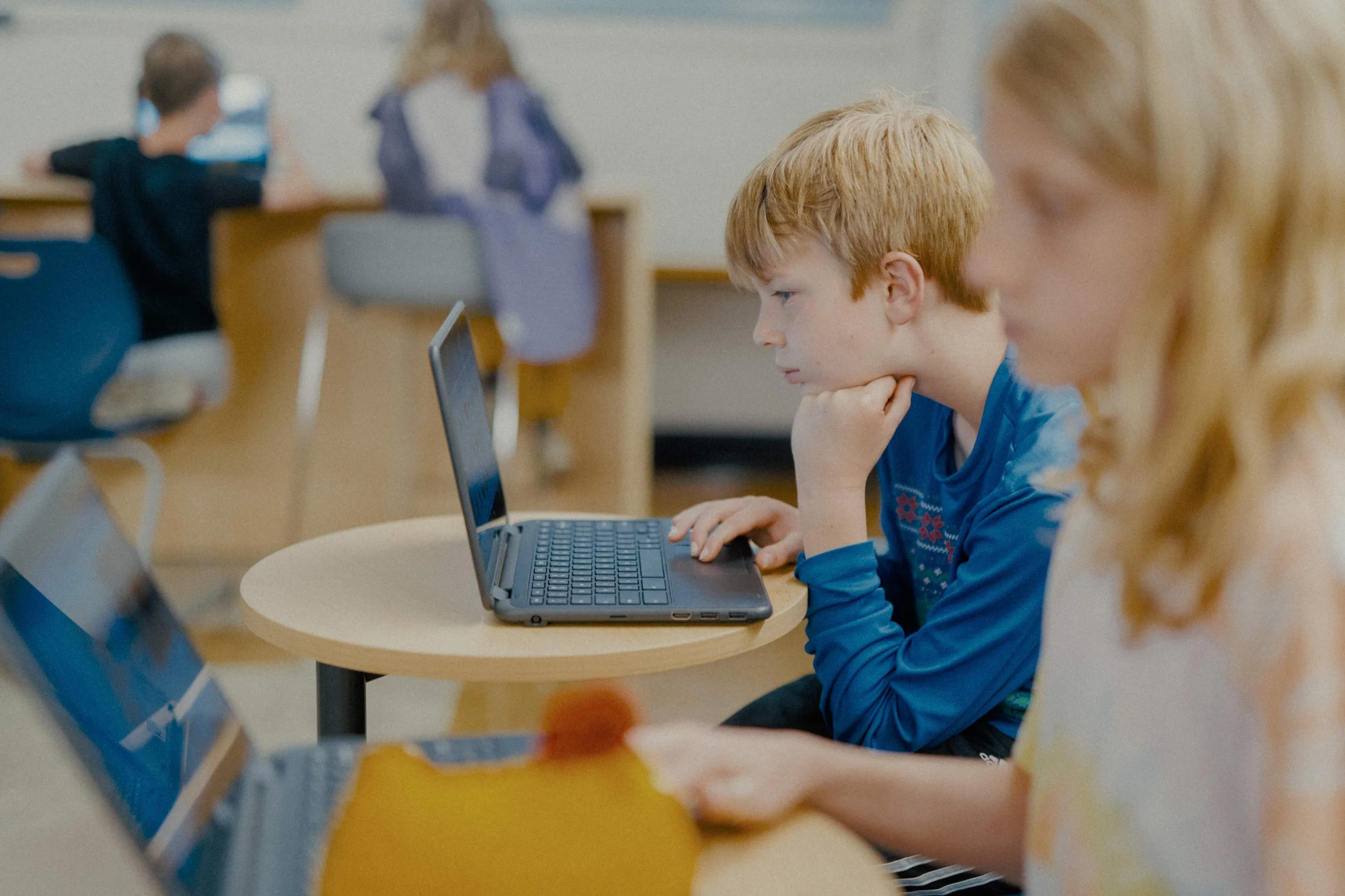 A young boy with red hair and a blue shirt sits at a desk, working on a laptop, with a focused expression. Next to him is a girl with blonde hair, also using a laptop. In the background, other children are seated at desks, some using computers.
