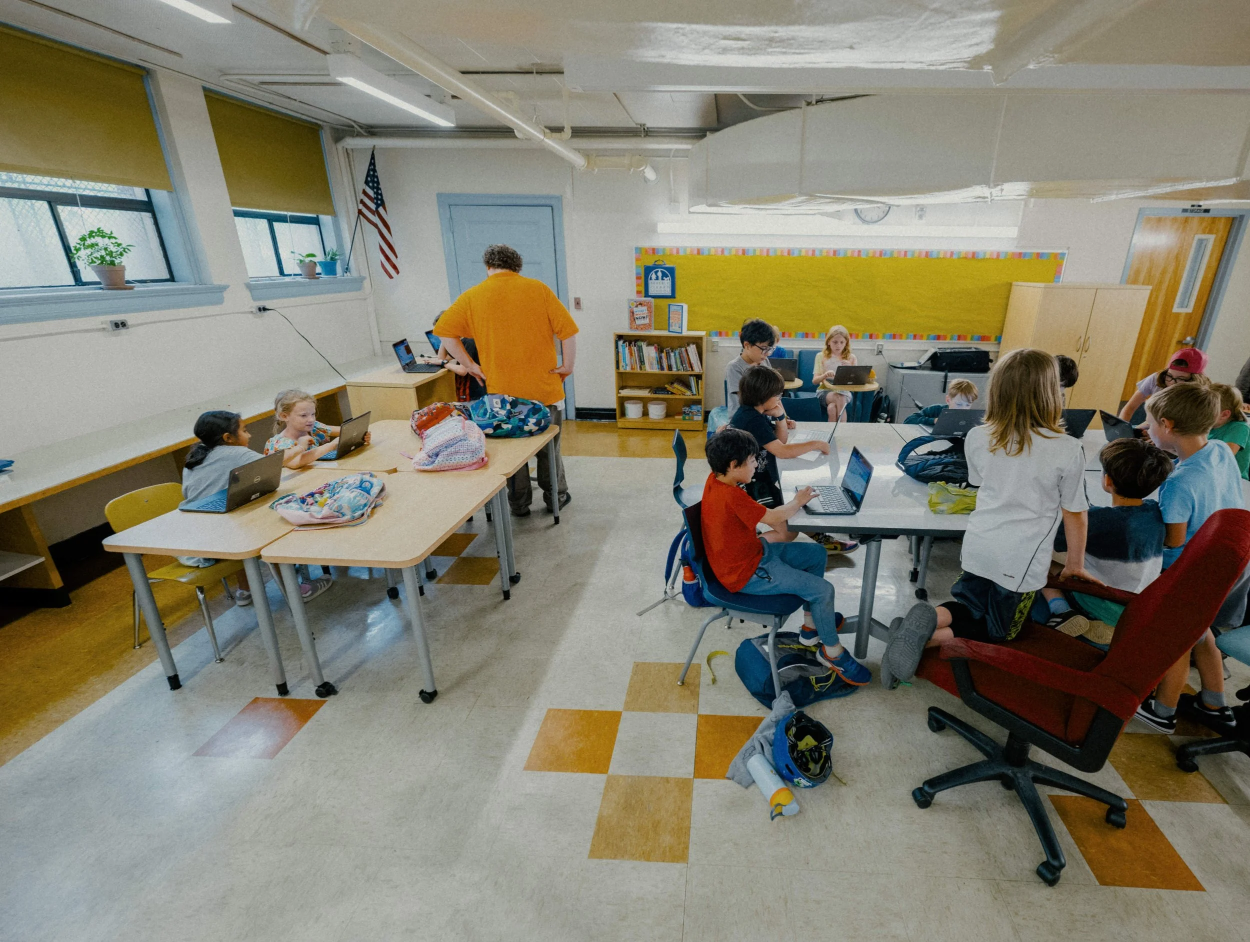 A classroom with children working on laptops and tablets, a teacher assisting students, and educational materials and books on shelves, with windows and potted plants in the background.