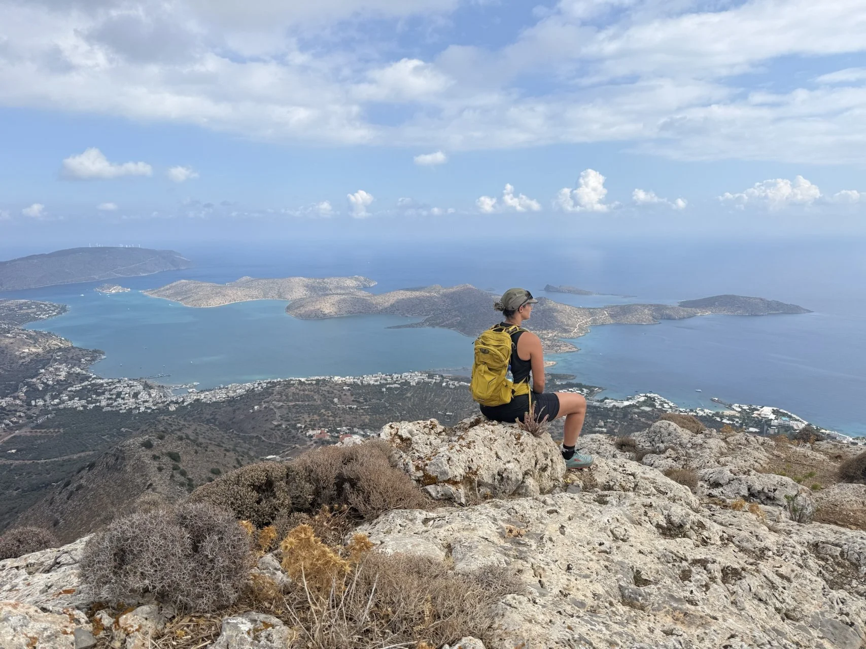 Een vrouw zit op een rots op een berg met een geel rugzak, kijkend naar het uitzicht op de zee en eilanden onder een bewolkte lucht.