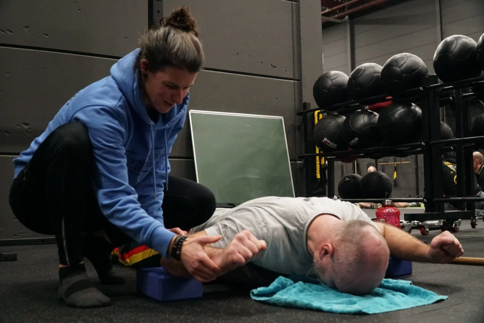 Persoon geeft eerste hulp aan een man die op de vloer ligt in sportschool met zwartwerpen, medicatie en een wit T-shirt.