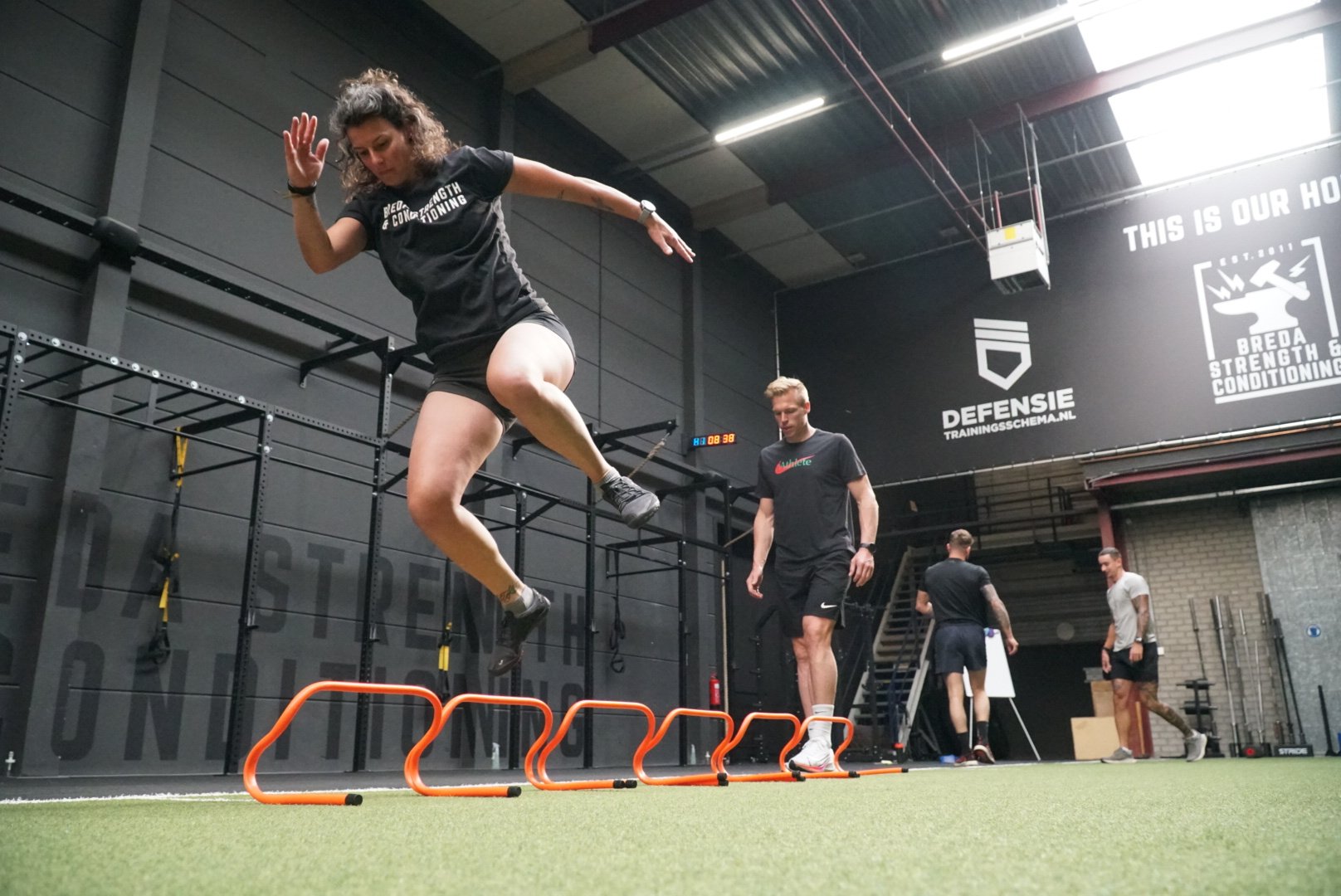Een vrouw springt over een hurdles tijdens een training, in een sportschool met andere personen die bezig zijn.
