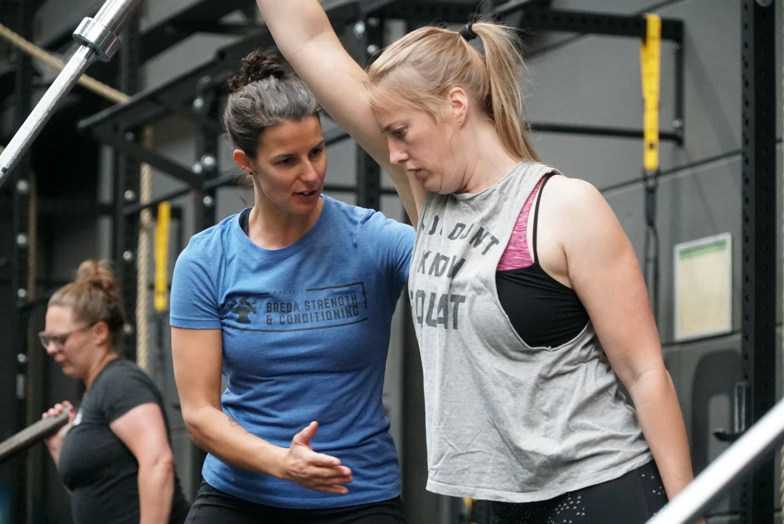 Een personal trainer helpt een vrouw bij een oefening in de sportschool, met andere mensen op de achtergrond die ook trainen.