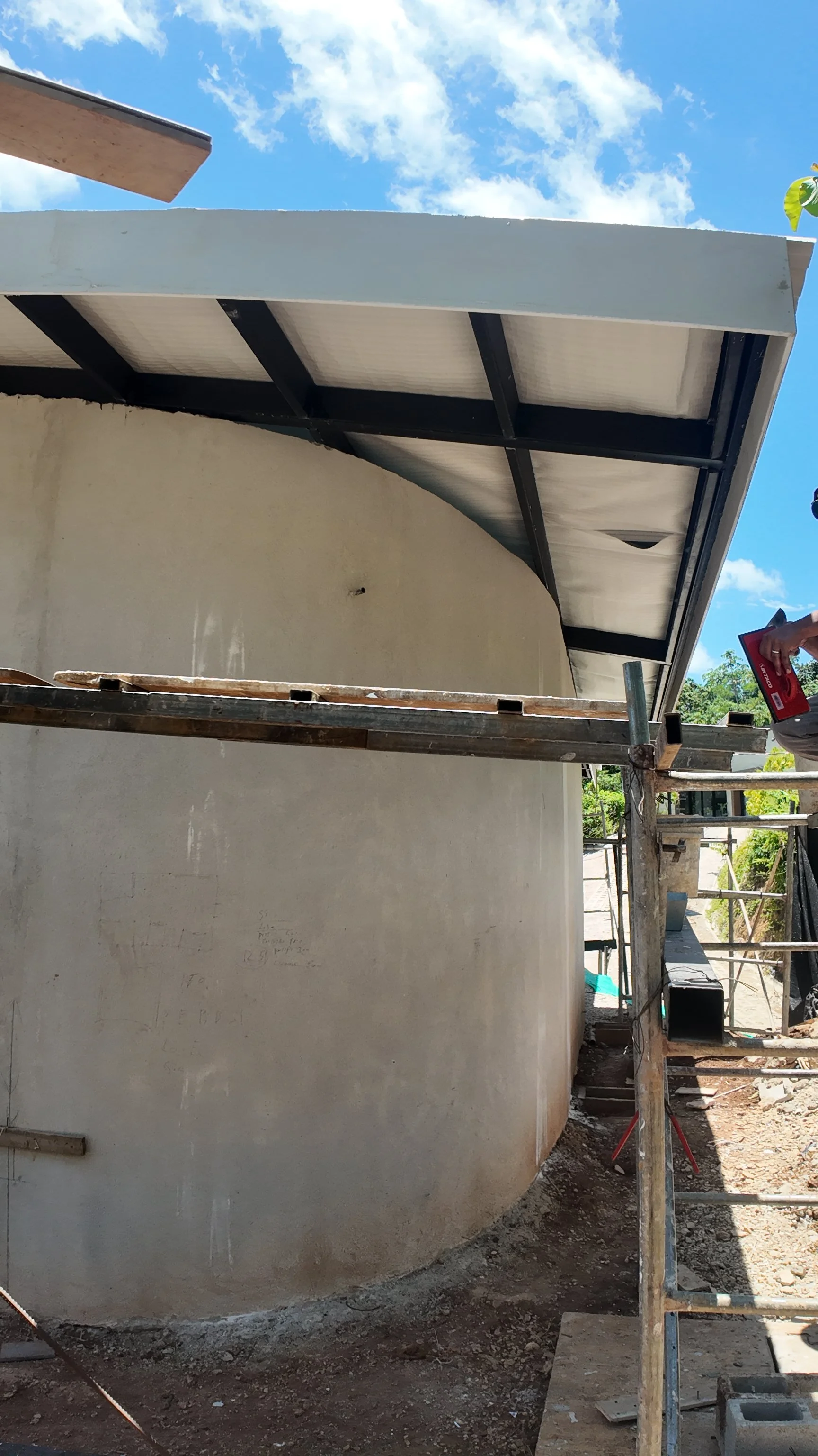 Construction site of a building with scaffolding and a roof extension, blue sky, and some greenery in the background.