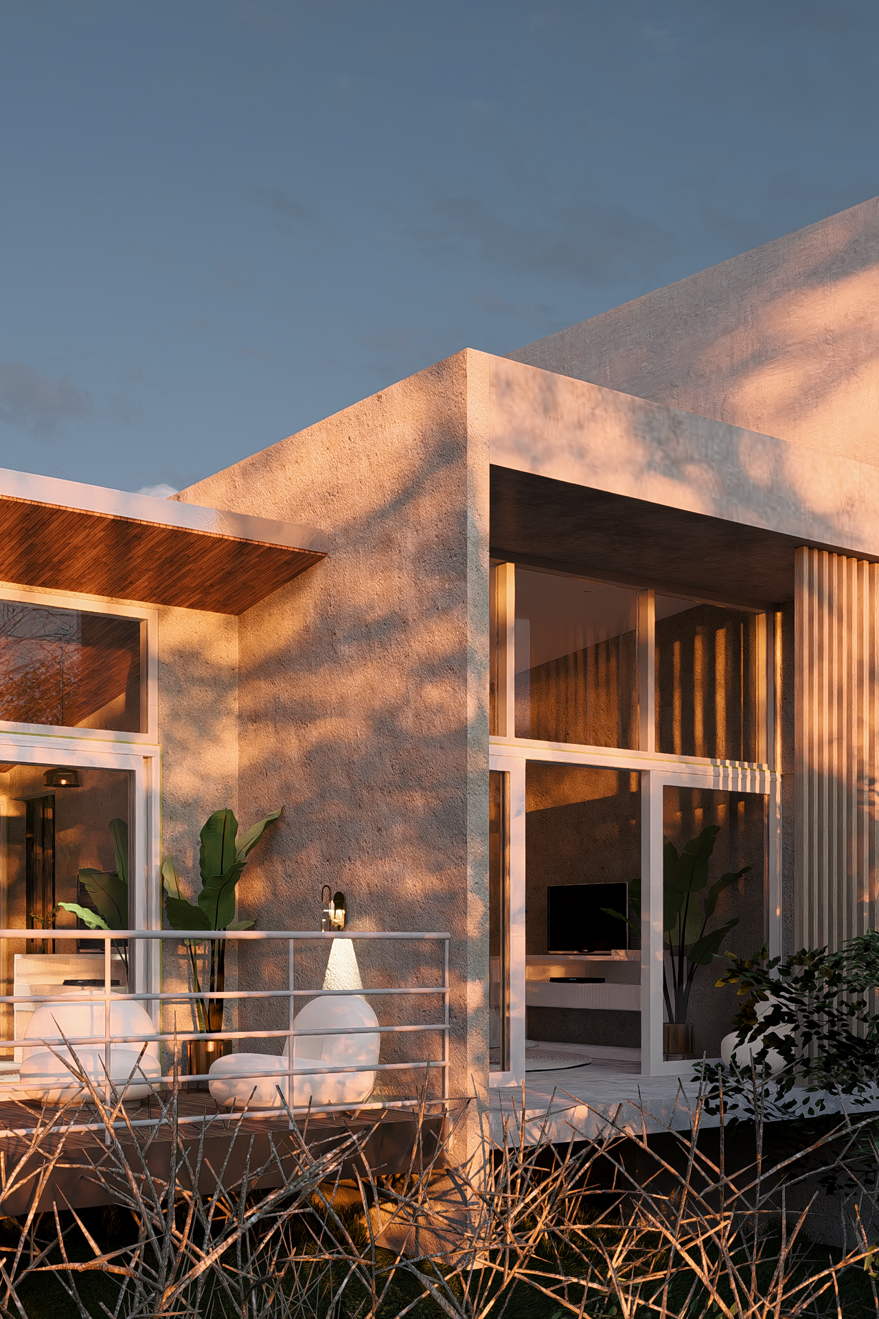 Modern residential house with large glass windows, textured concrete walls, and a wooden ceiling on the porch, illuminated by warm sunlight, with plants and dried branches in the foreground and a clear blue sky in the background.