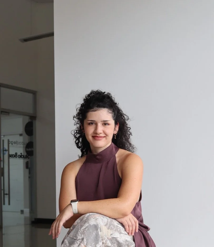 A woman with curly dark hair sitting against a plain white wall in a modern indoor setting, wearing a sleeveless burgundy top, patterned pants, and a white smartwatch, smiling at the camera.