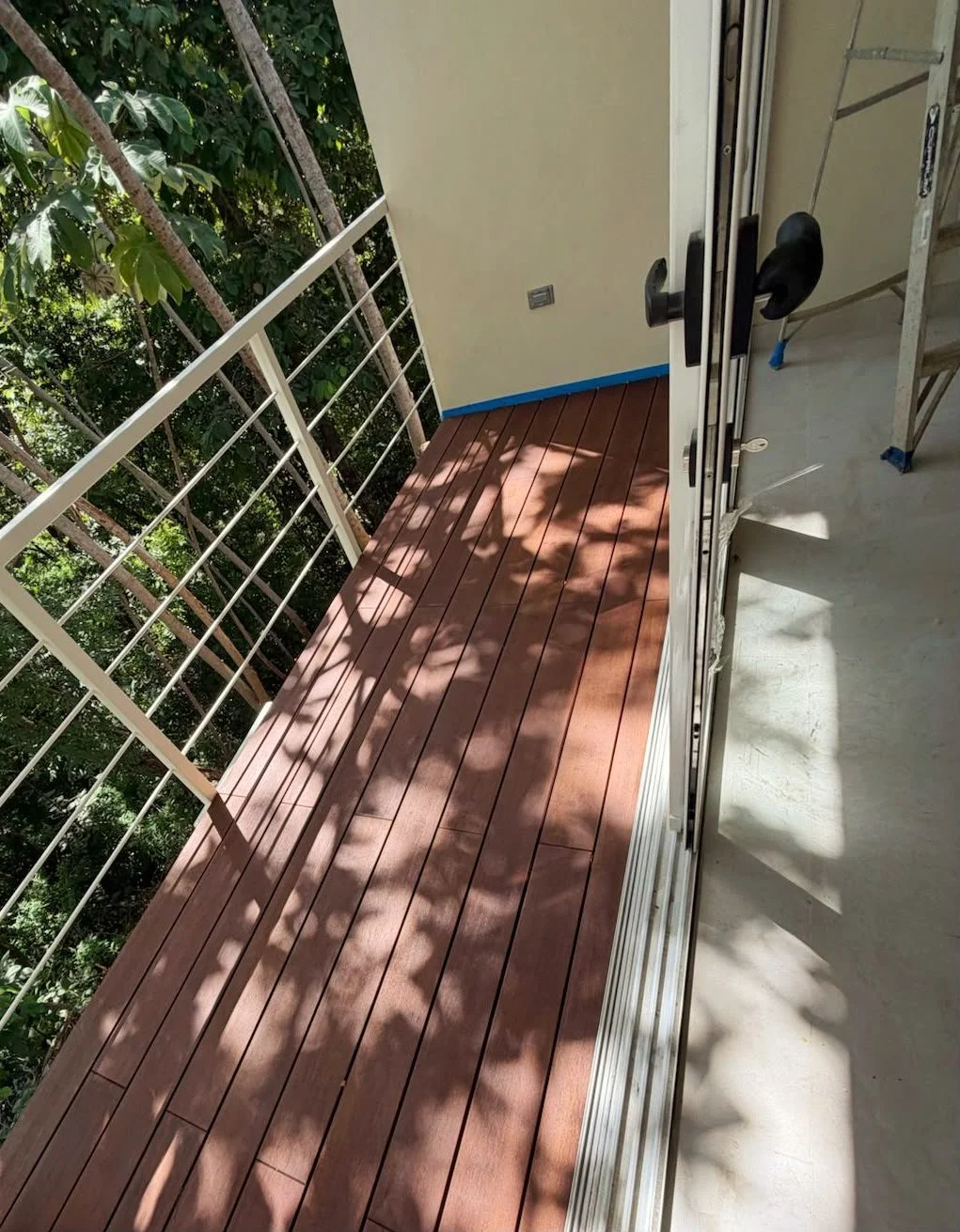 Small balcony with wooden floor and white metal railing, overlooking a lush green area with trees. Part of an open sliding glass door is visible.