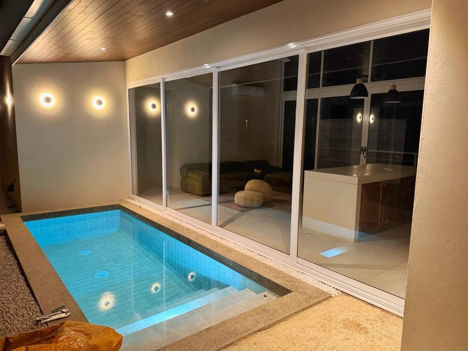 Indoor pool area with a small illuminated blue pool, sliding glass doors in the background, and modern interior decor with warm lighting.
