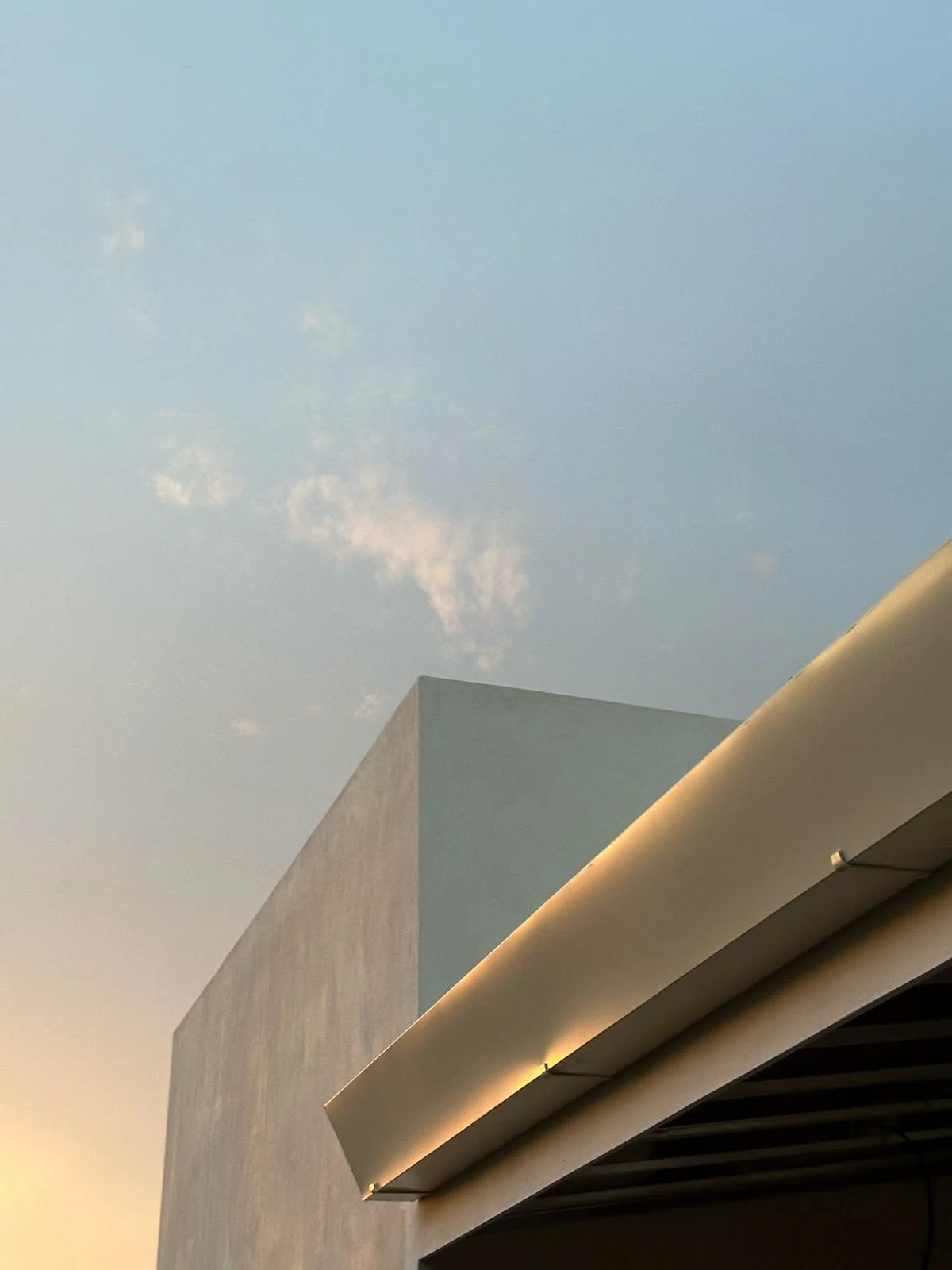 A modern building with a minimalist design featuring a light-colored facade and an overhanging roof with embedded lighting, against a partly cloudy sky at sunset.