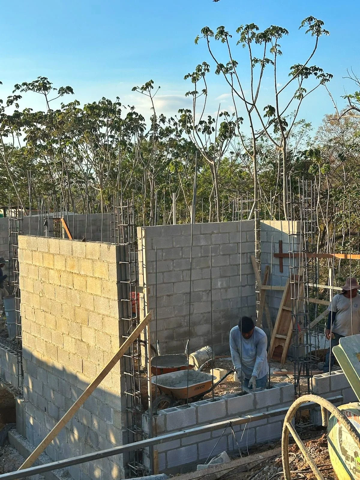 Construction workers building a brick wall on a house under construction, with steel rebar and wooden supports, surrounded by trees under a blue sky.