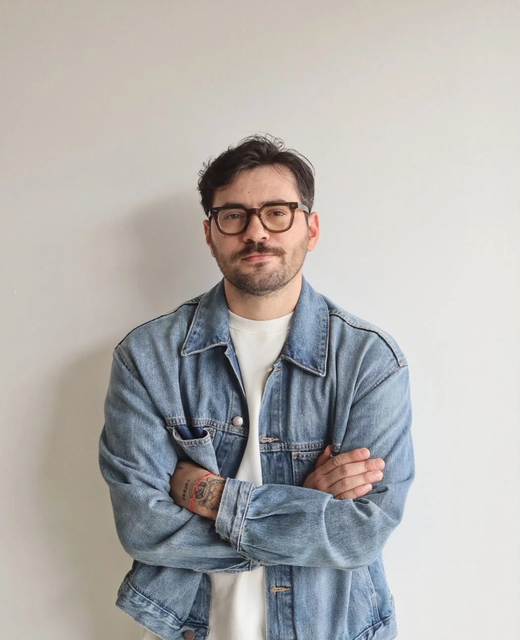A man with dark hair, glasses, and a beard, standing with arms crossed against a plain white wall. He is wearing a denim jacket over a white shirt.