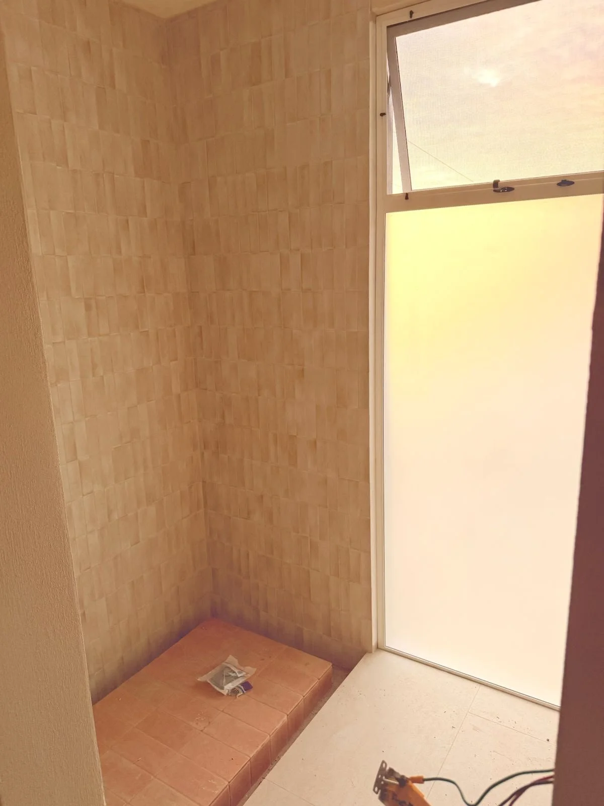 Empty indoor space with beige tiled wall and a window with frosted glass, a small tiled platform in the corner, and some tools on the floor.