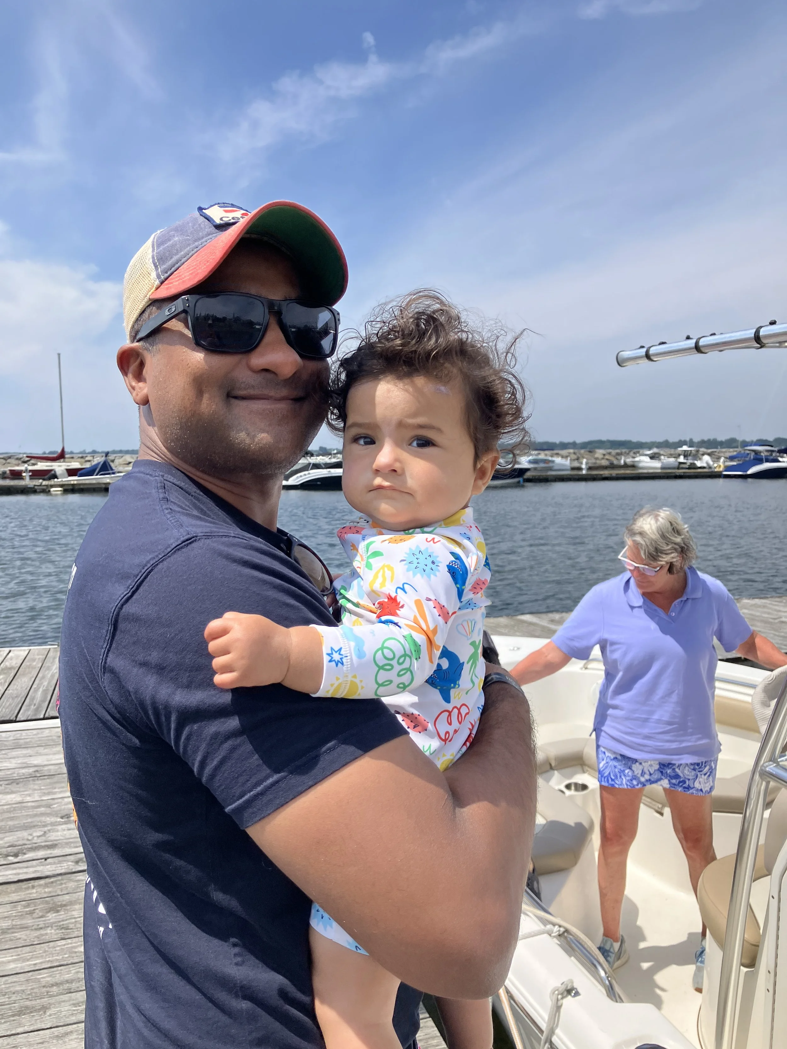 A man wearing sunglasses and a cap holding a young child while standing on a dock near boats in a marina, with an older woman in the background.