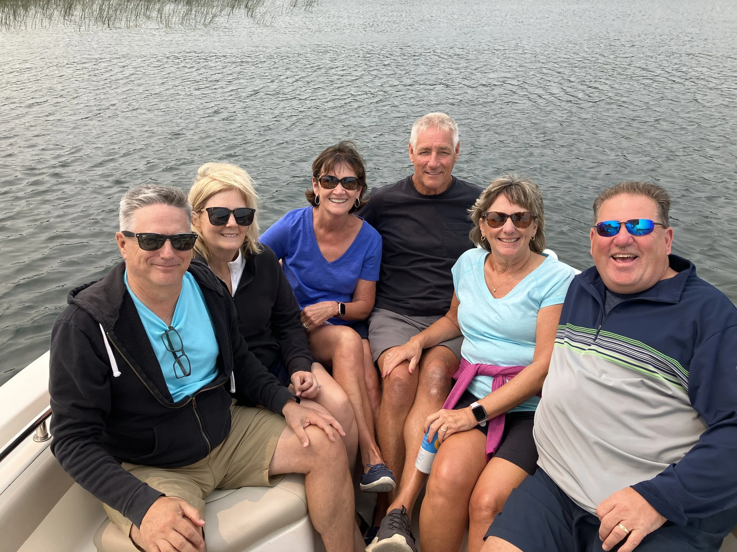 Group of six people sitting on a boat, smiling, with water in the background, wearing sunglasses and casual clothing.