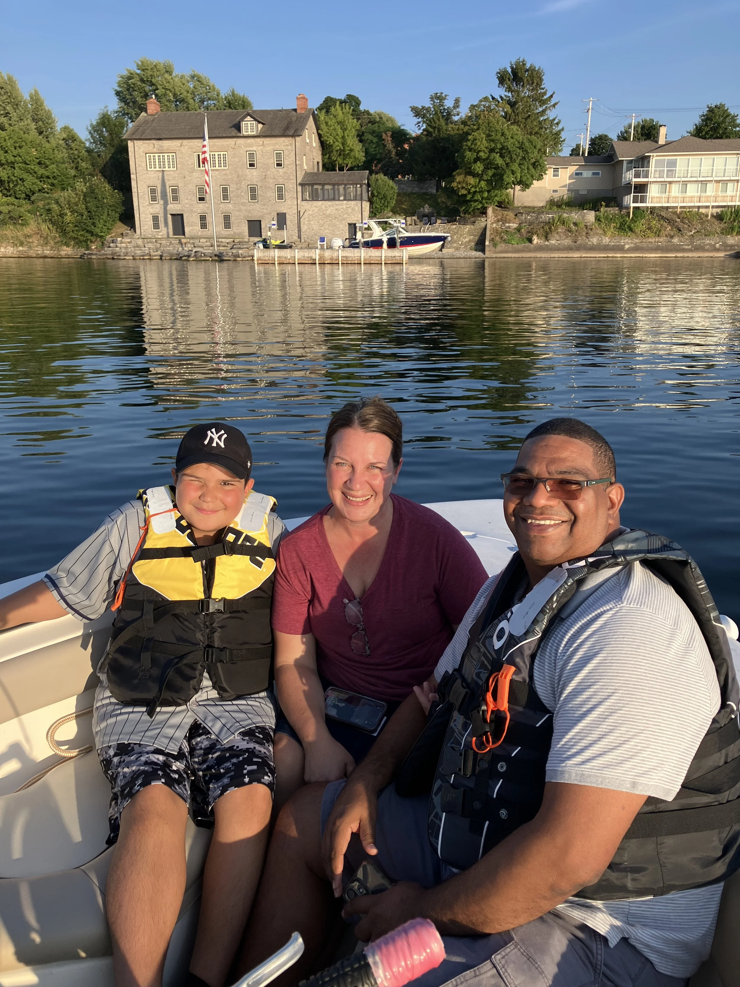 A family of three on a boat smiling for a photo with houses, trees, and water in the background.