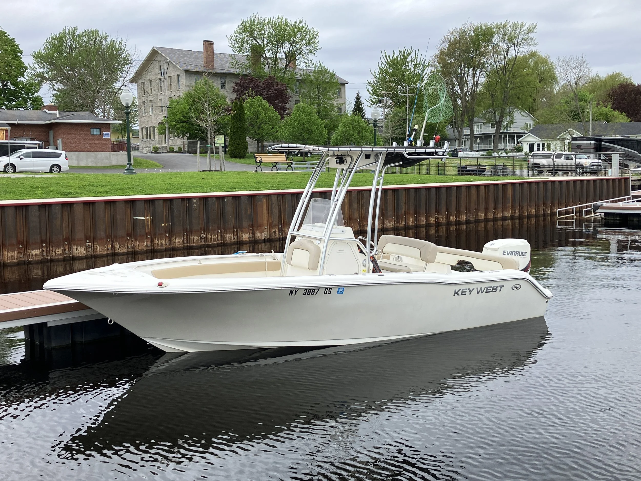 A white Key West boat docked at a marina with a rural residential neighborhood in the background, featuring trees, houses, and parked cars.