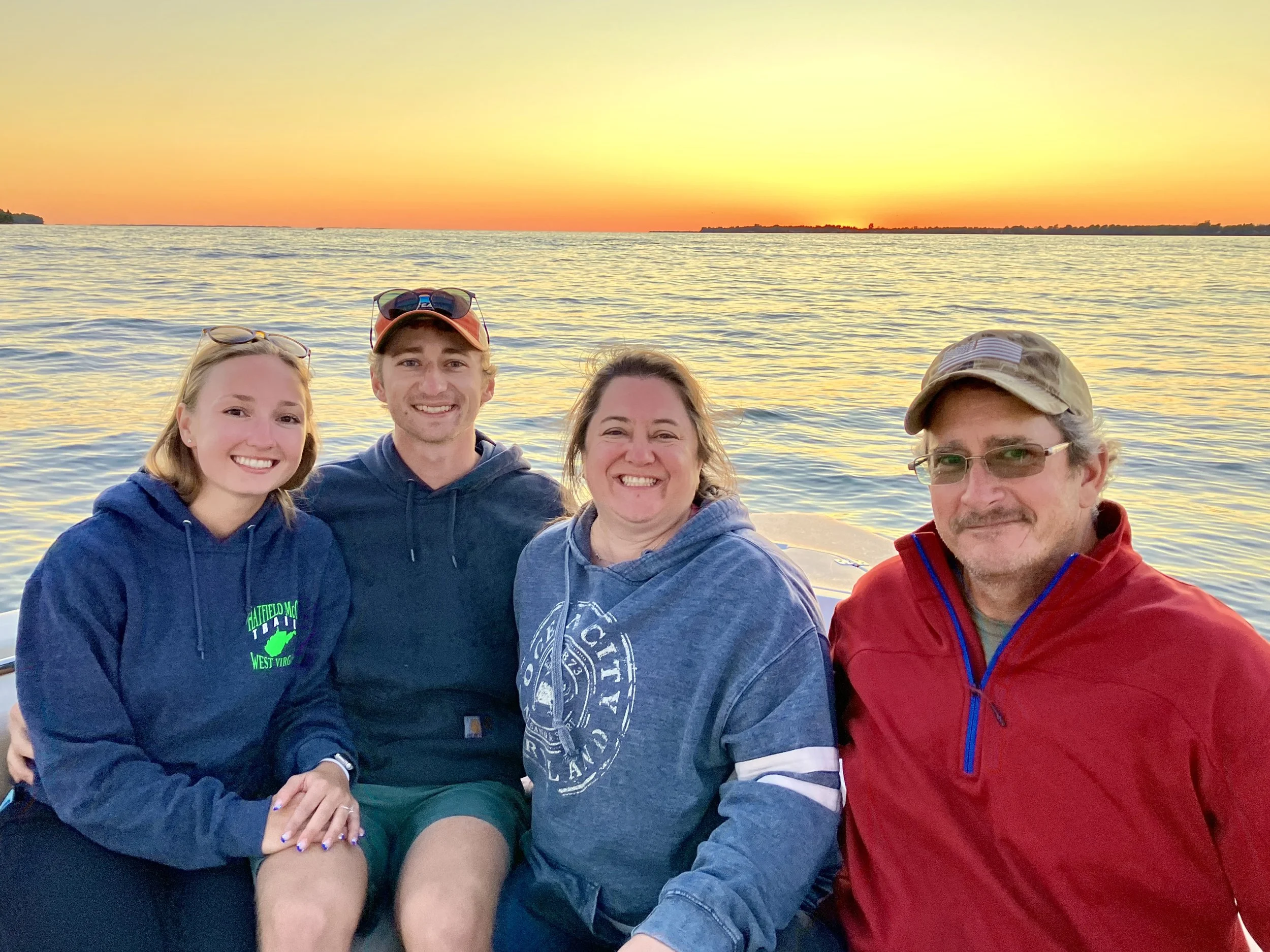 A group of four people sitting on a boat at sunset on a body of water. They are smiling and wearing casual, warm clothing. The sunset creates a colorful sky with shades of orange, yellow, and pink.