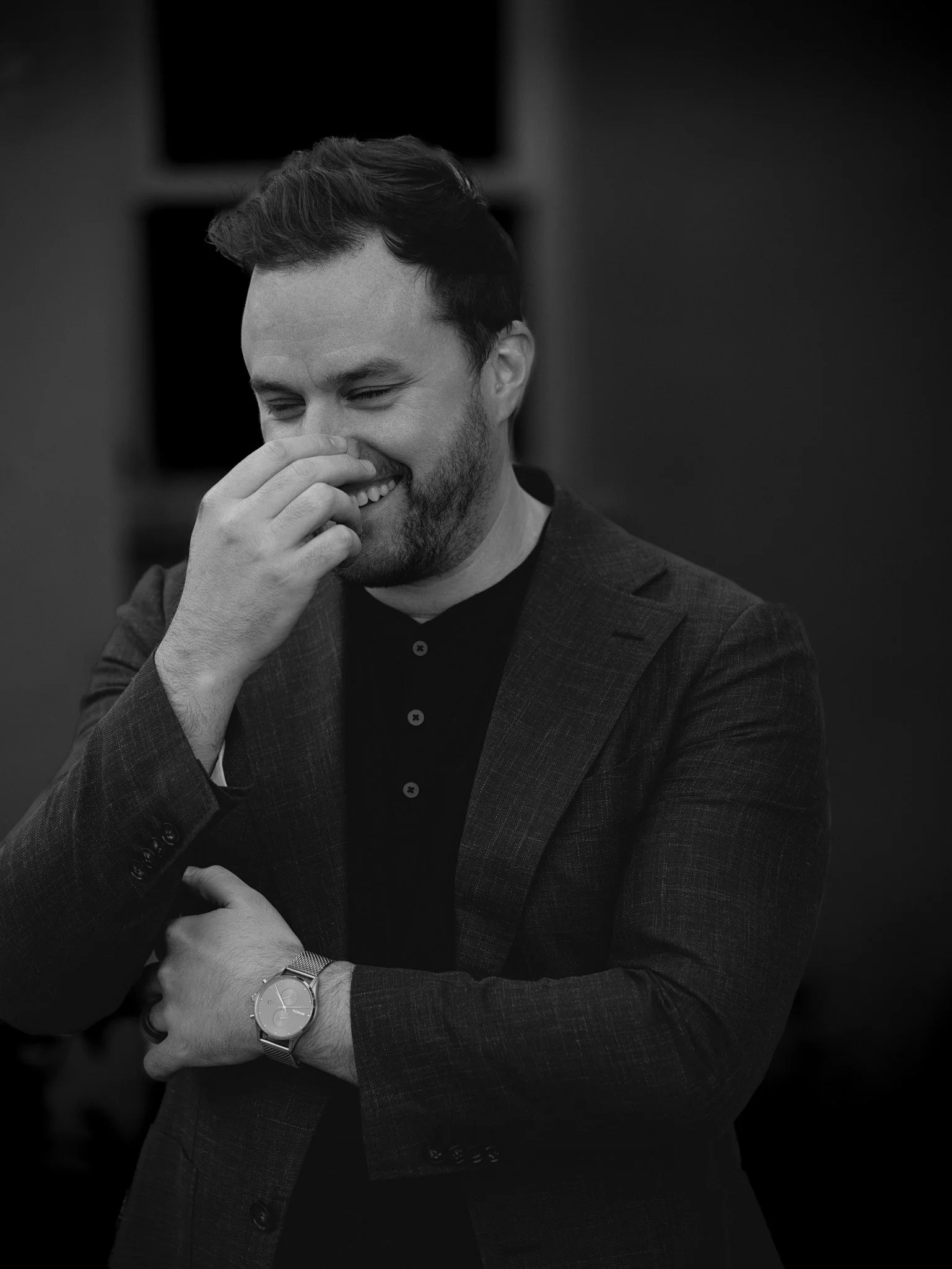 Black and white photo of a man smiling, wearing a suit, holding his nose with one hand and crossing arms.