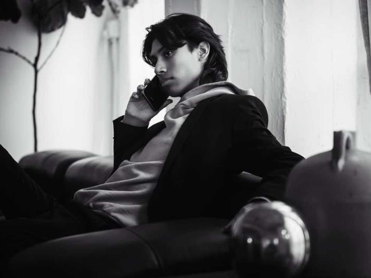 Person sitting on a couch talking on a smartphone, wearing a jacket over a hoodie, in a black and white setting.