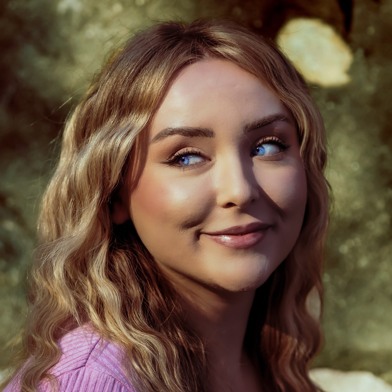 Smiling woman with long blonde hair and blue eyes wearing a purple top, looking to the side, outdoors.