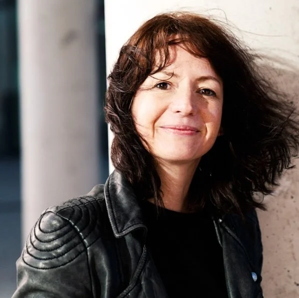 Wissenschaftsjournalistin Monika Sax in schwarzer Lederjacke lehnt sich an eine Wand, mit halblangen, dunklen Haaren und einem Lächeln.
