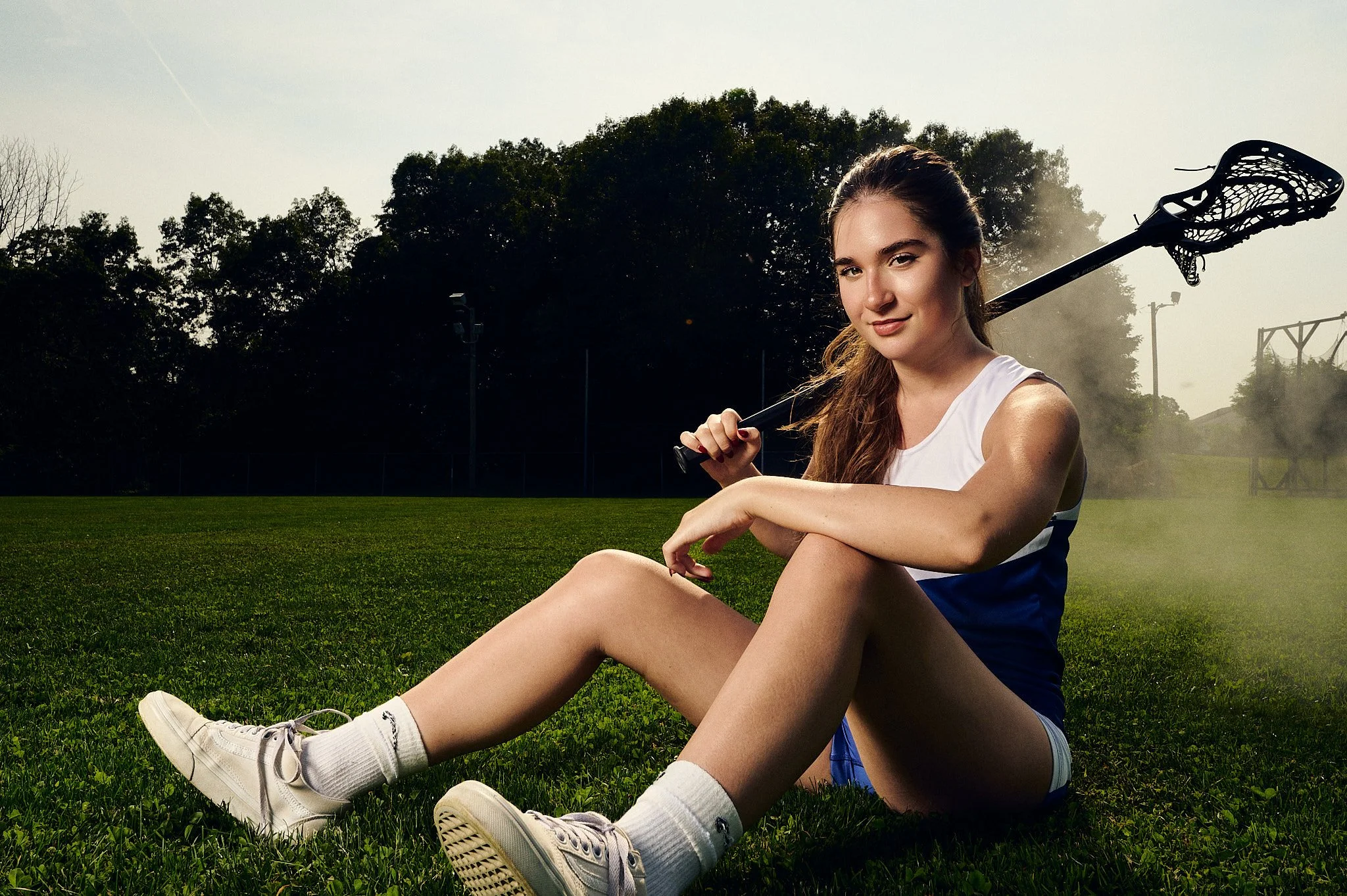 Young woman in lacrosse uniform sitting on field with stick over shoulder, natural light portrait, Manchester CT