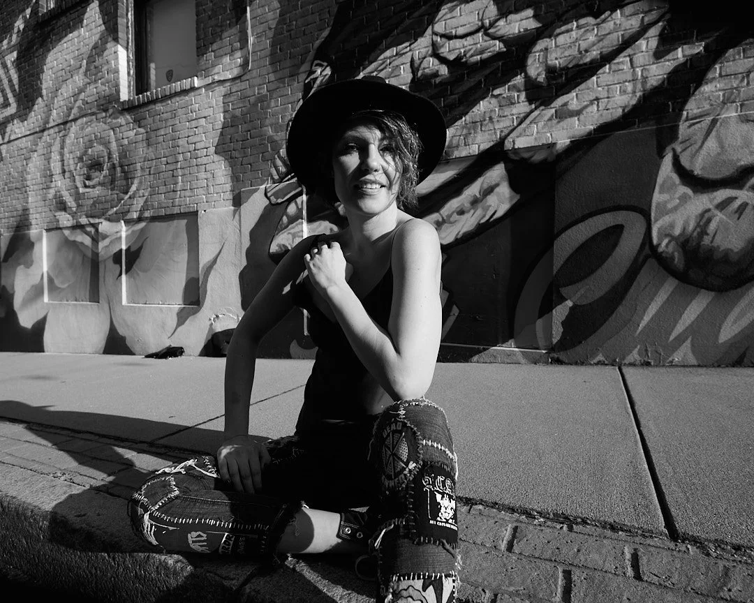Rick Morgan Photography Manchester CT. Person sitting on sidewalk in front of mural, wearing a hat and patterned pants, smiling, black and white photo.