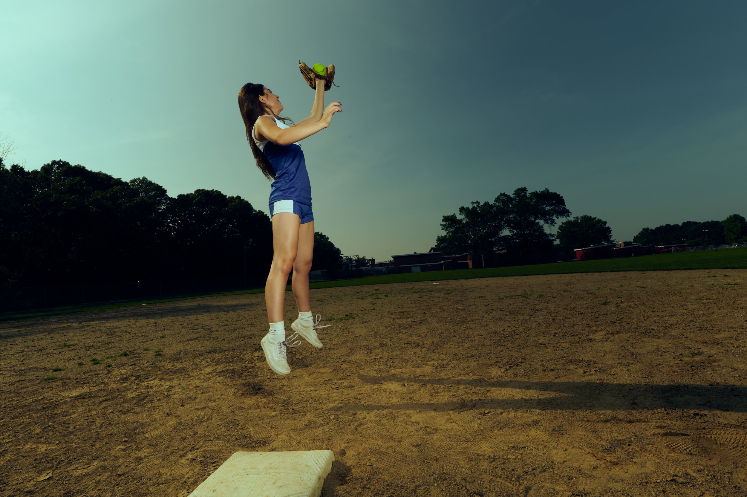 High School Senior Photography Manchester CT. and surrounding area. Young woman in athletic uniform jumping to catch a baseball with glove during daytime on baseball field.