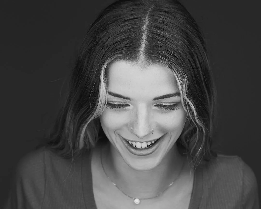 Rick Morgan Photography Manchester CT. Black and white portrait of a smiling person with wavy hair and a nose piercing, looking downwards.