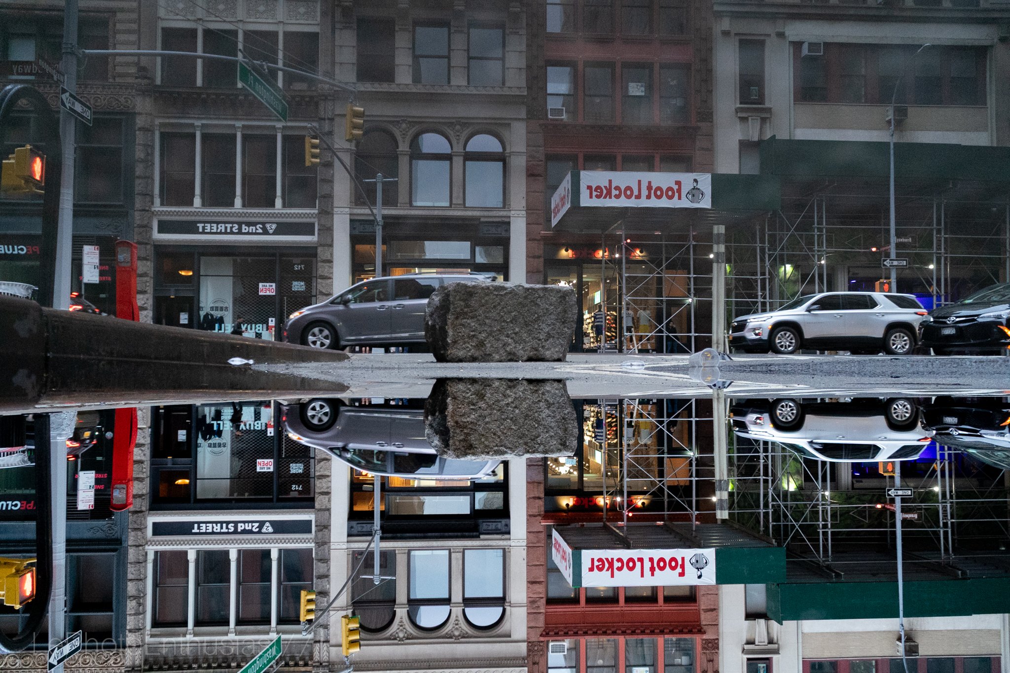 City street scene with parked cars, buildings with scaffolding, a Food Locker sign, and a large rock on a reflective surface creating a mirror image.